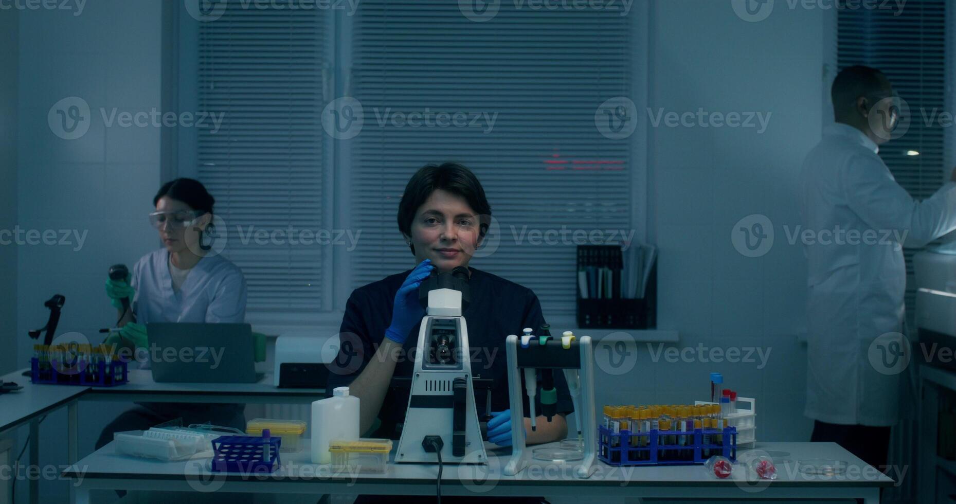 A young woman in a lab coat smiles at the camera sitting in front of a microscope in the laboratory, working next to her colleagues photo