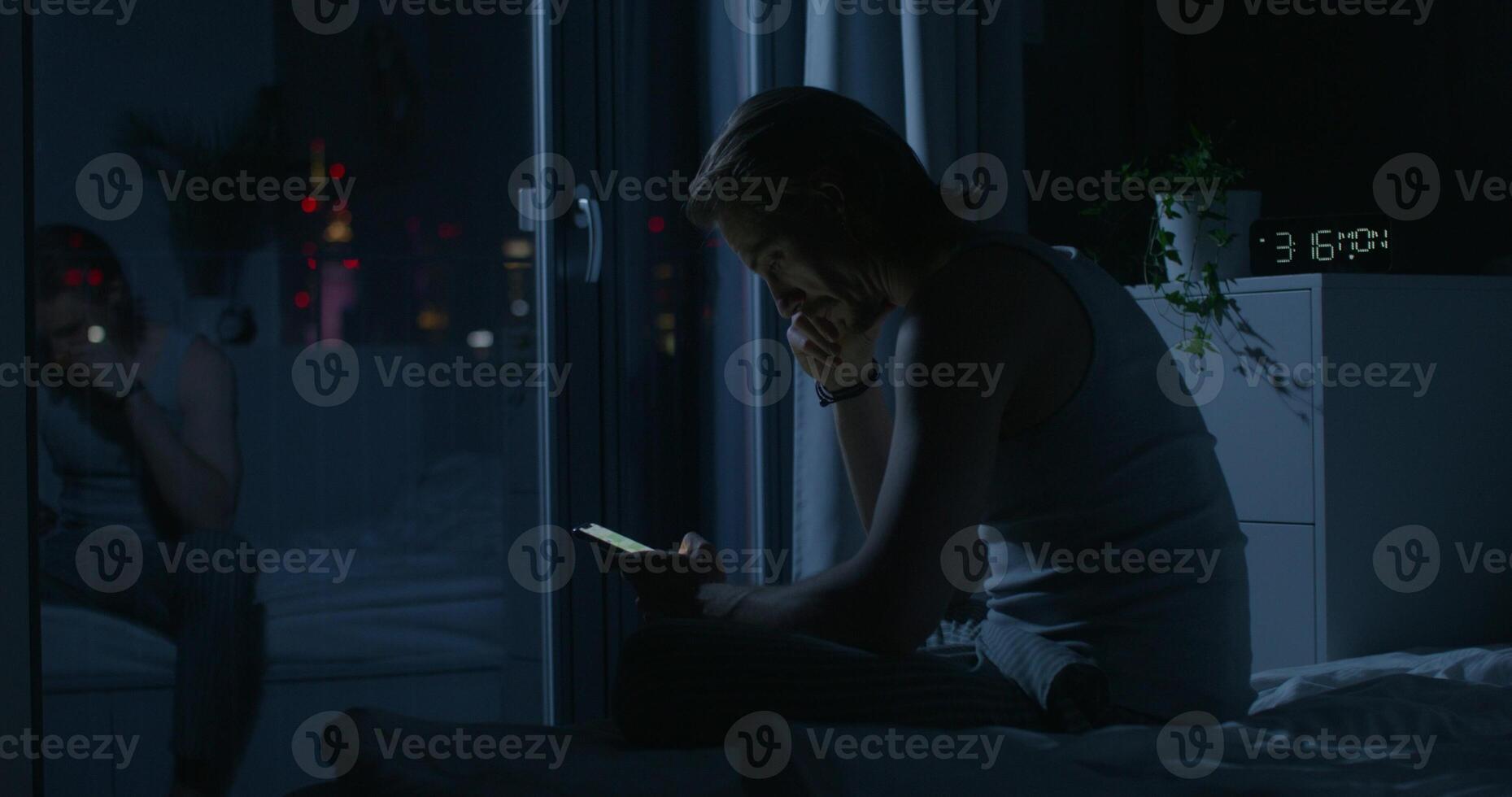 A man in pajama pants uses a mobile phone while sitting on the bed at night. Dark room, home interior - interior photo