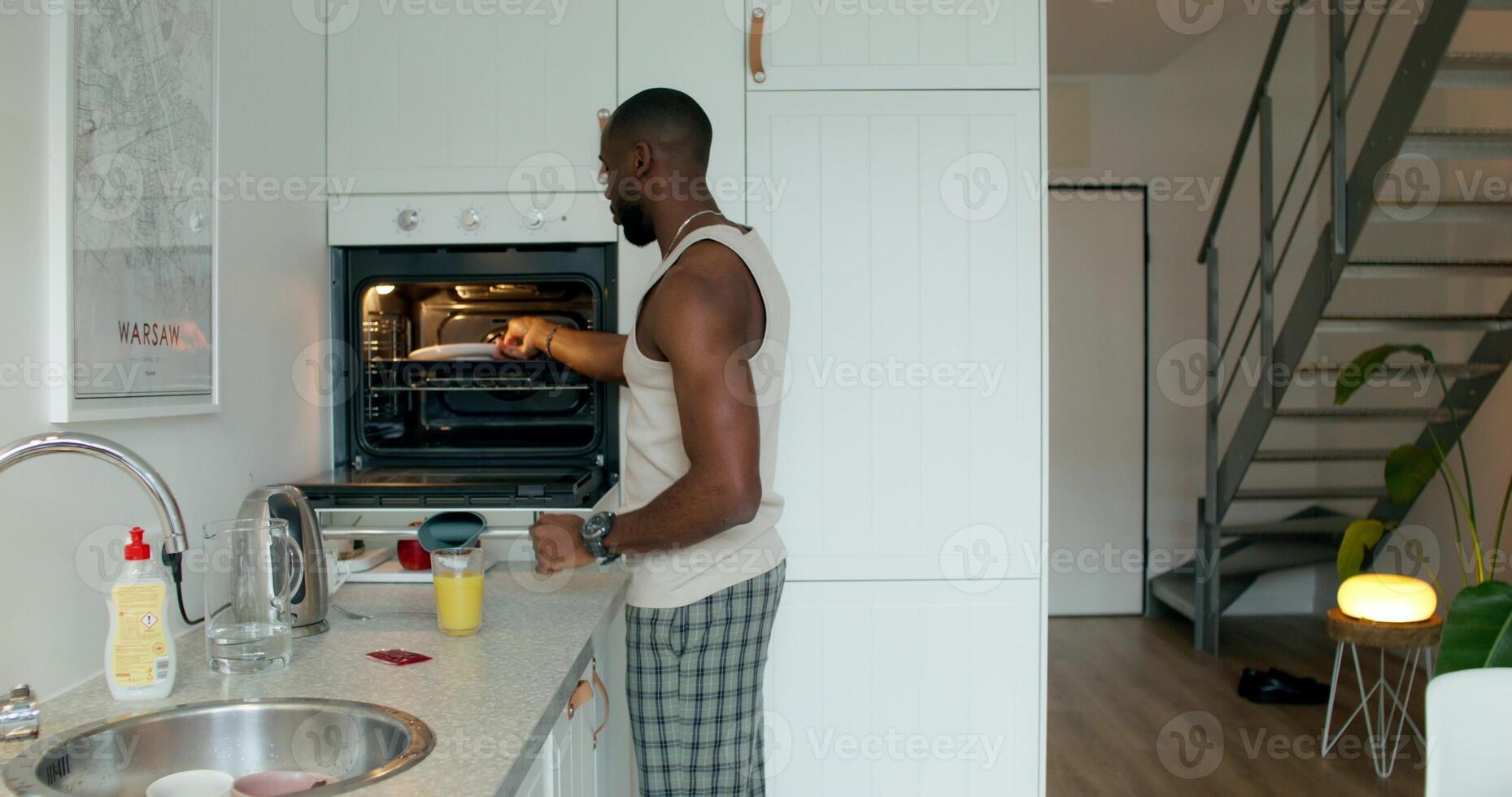 A man in pajamas takes the ready-made food out of the oven and sits down at the table to eat. Home interior, daylight photo