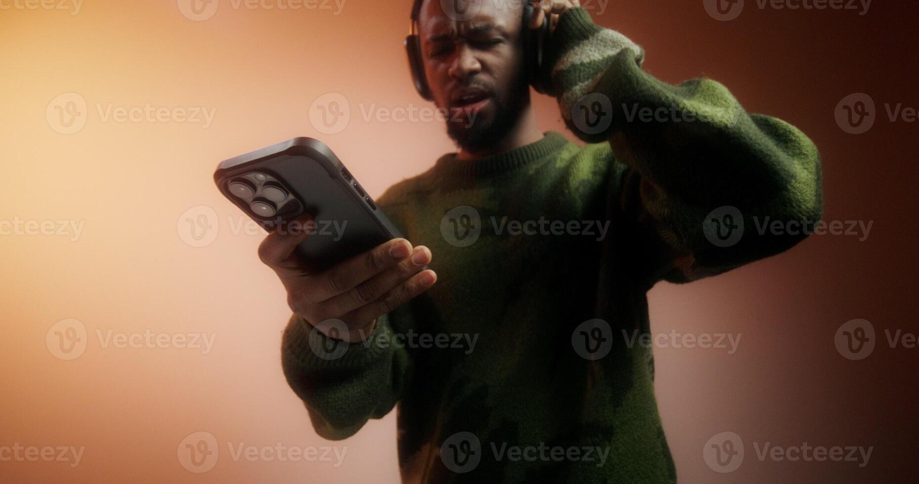 A bearded man in a sweater and headphones turns on music on his mobile phone and starts dancing standing on an orange background in the studio photo