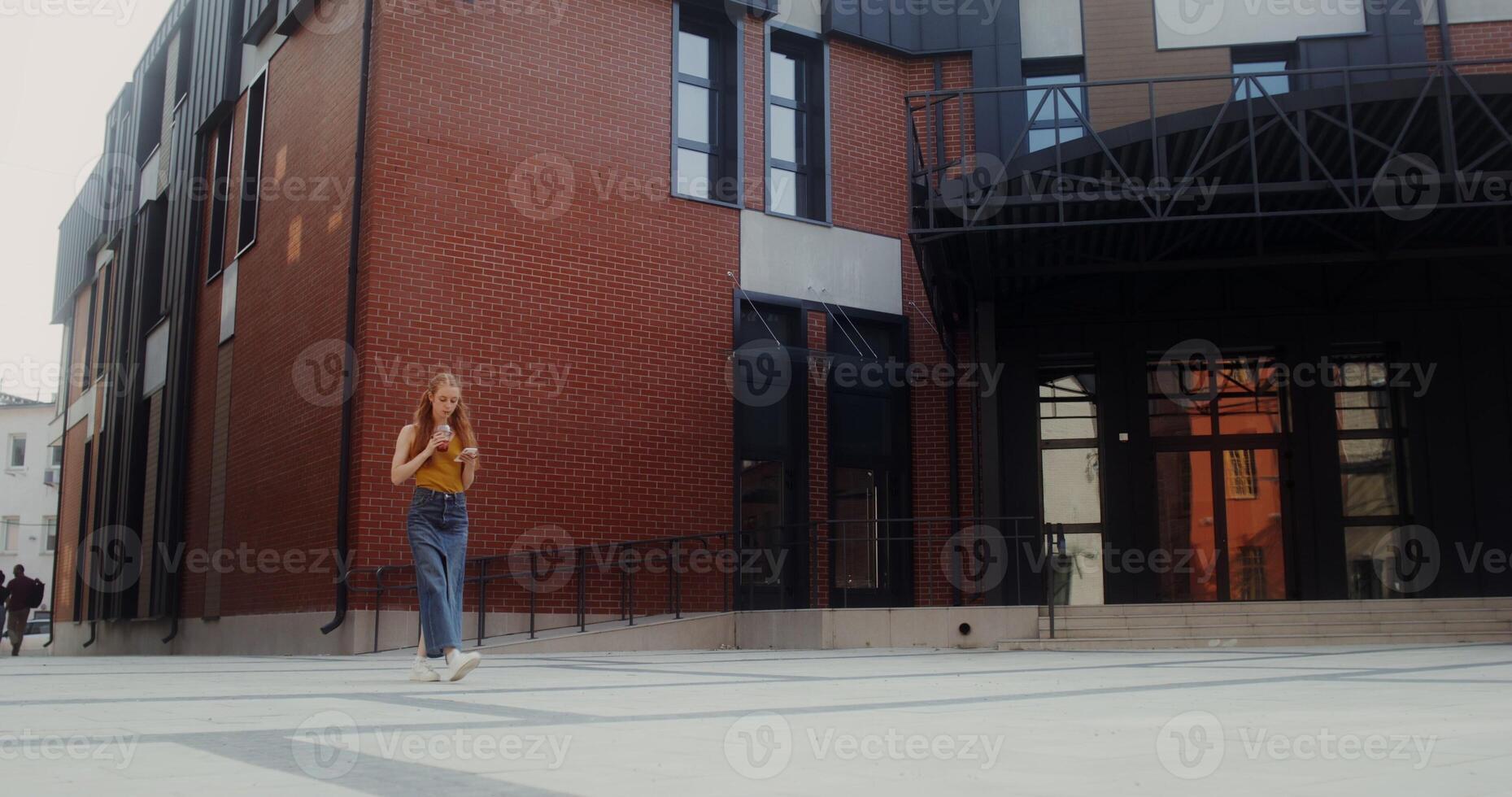 A young red-haired woman uses a mobile phone and drinks juice from a disposable cup with a straw while walking on a city street - disposable photo