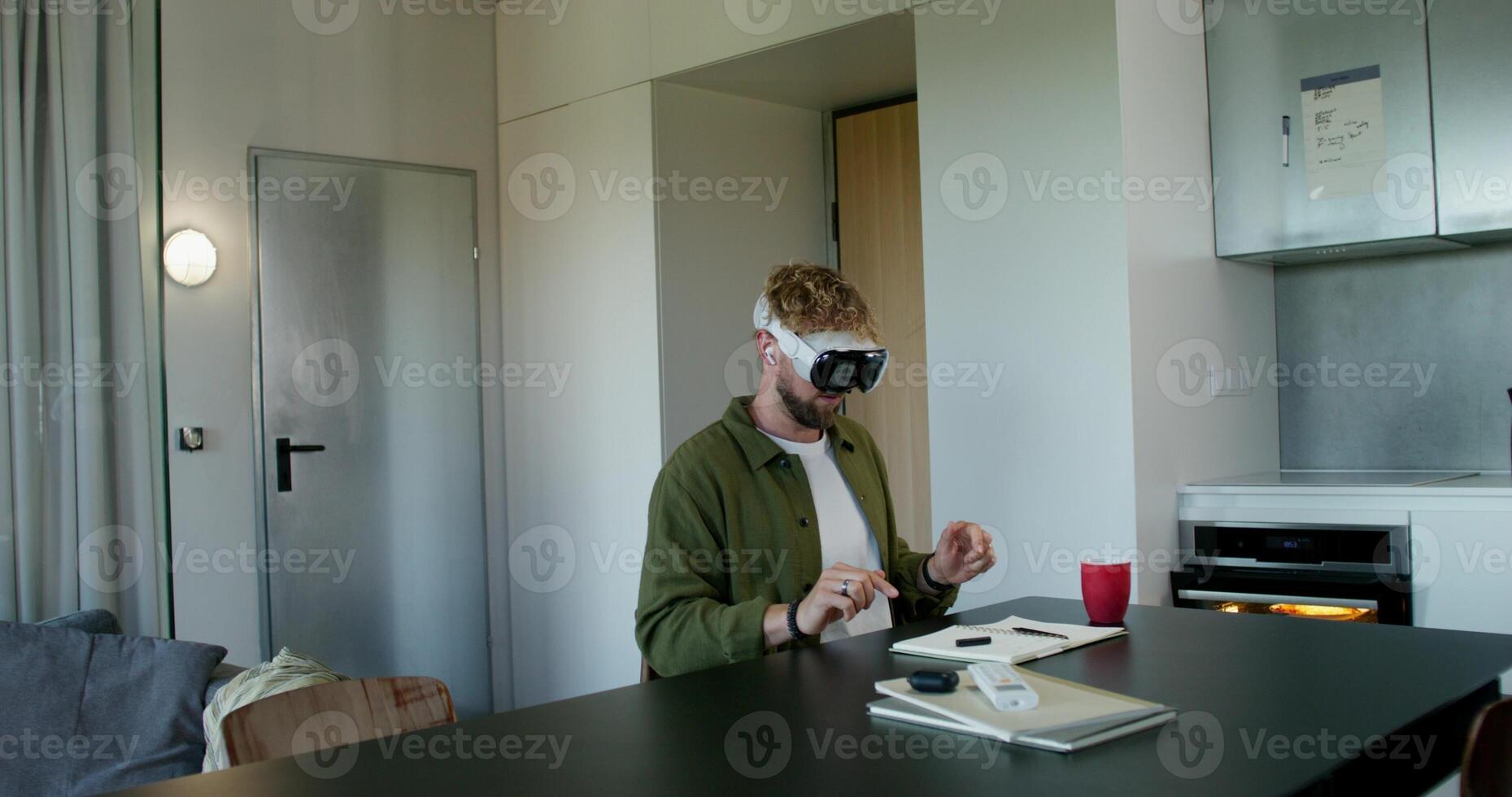 A man in a virtual reality headset typing on a virtual keyboard sitting at a table at home. Daylight photo