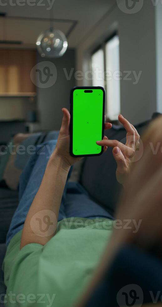 A woman uses a mobile phone with green screen lying on the couch at home. A rear view of her hands, an unrecognizable person - unrecognizable photo