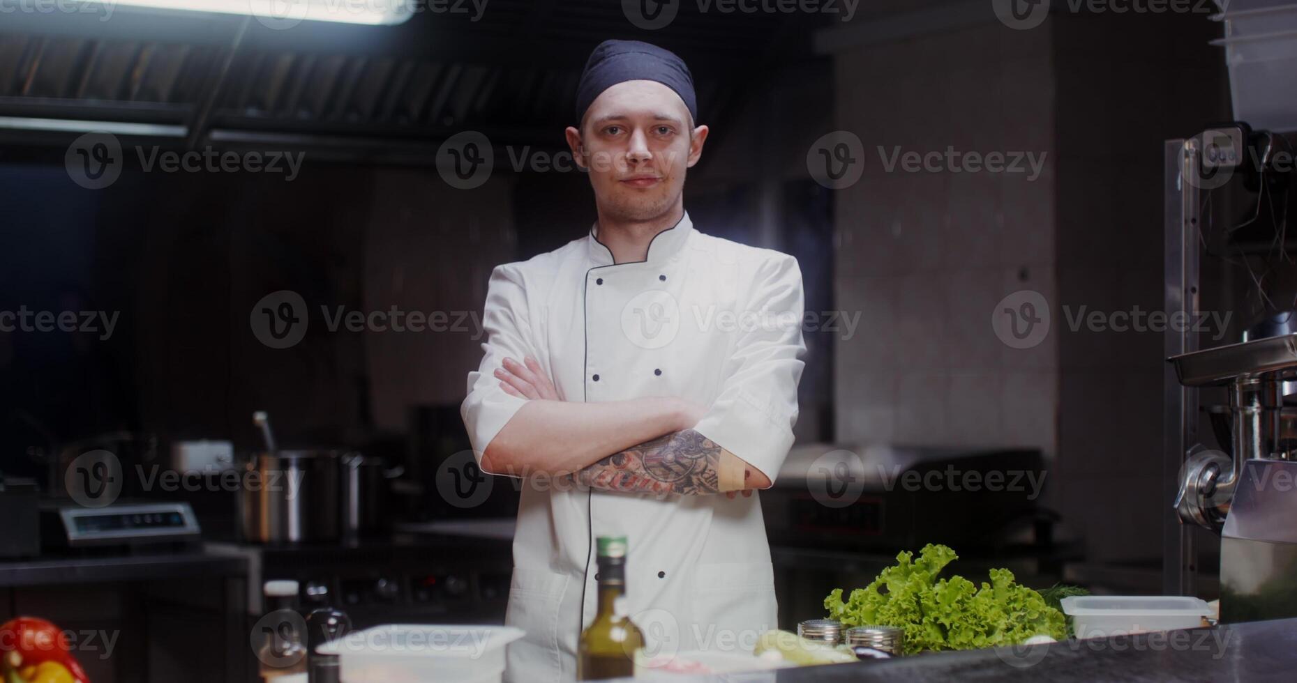 A man in a chef's uniform with a headband looks at the camera with a slight smile, standing in a modern kitchen with his arms crossed at his chest photo