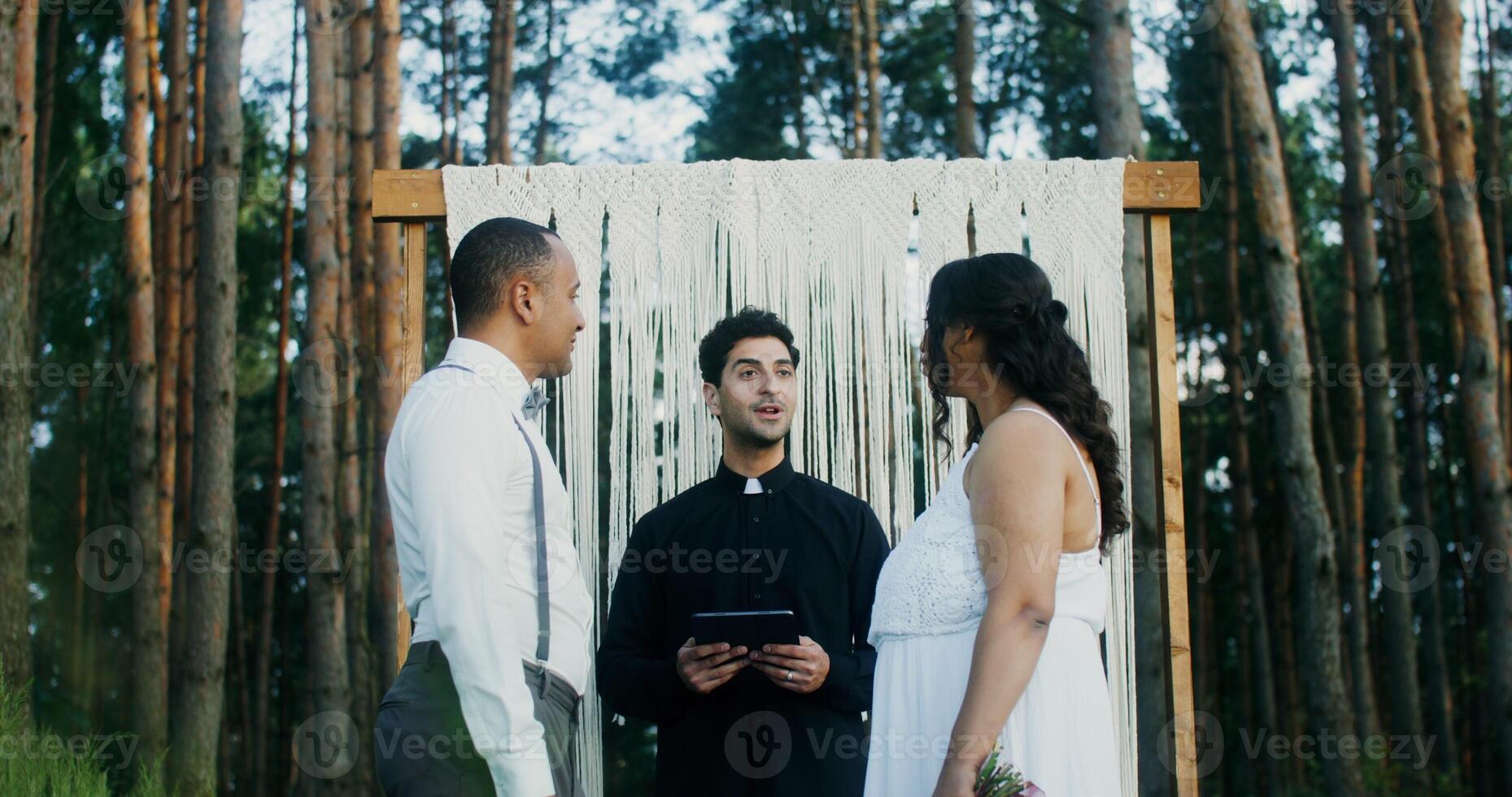 The bride and groom kiss at the wedding standing in front of the altar. Wedding in the forest with a priest - Wedding photo