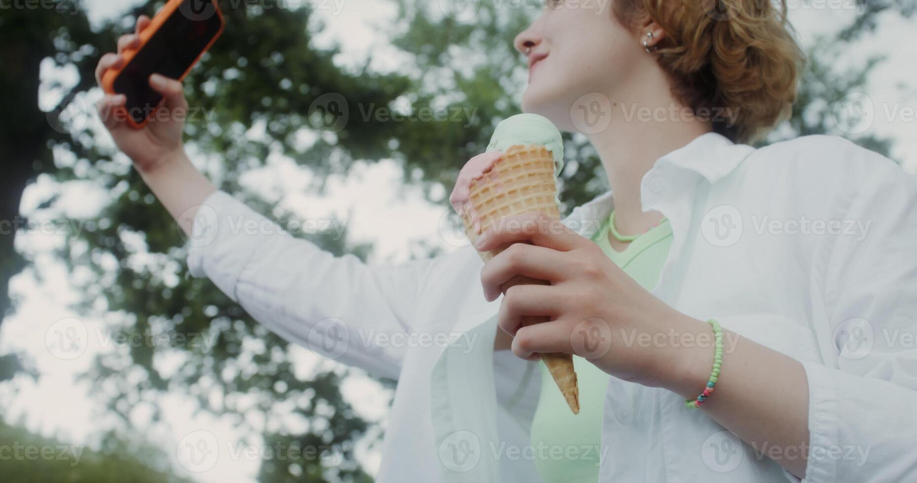 A young woman takes a selfie on a mobile phone standing in a park with ice cream in her hand photo