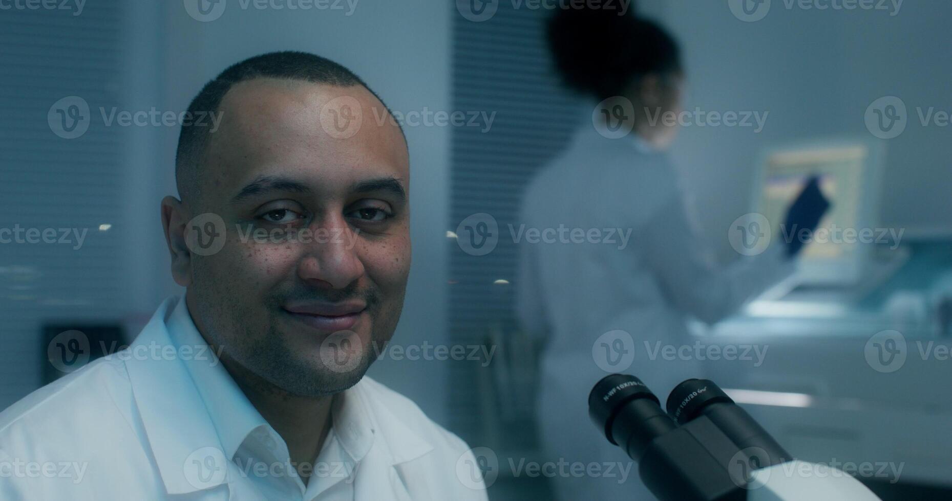 A man in a lab coat looks at the camera sitting in front of a microscope in a medical laboratory - medical photo