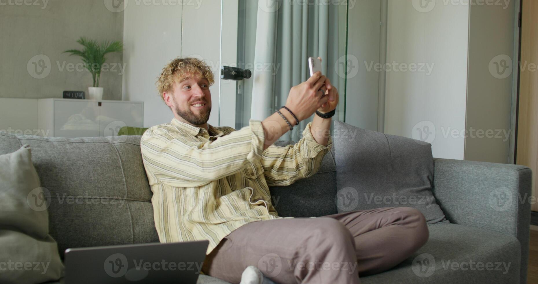 A bearded man in casual clothes is talking on a chat on a mobile phone sitting on the couch at home. The laptop is next to him. Daylight - clothes photo