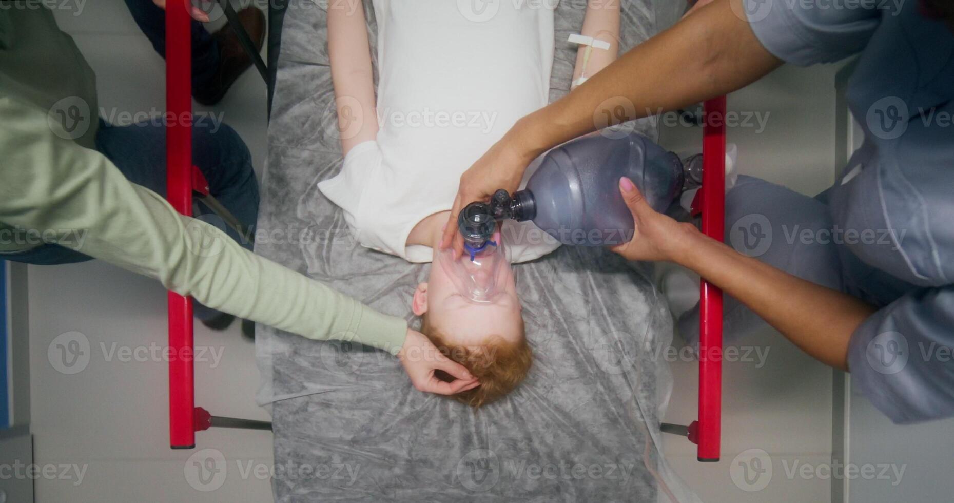 Medical staff and parents carry the boy on a gurney, giving him oxygen with a mask, an overhead view of his face - giving photo