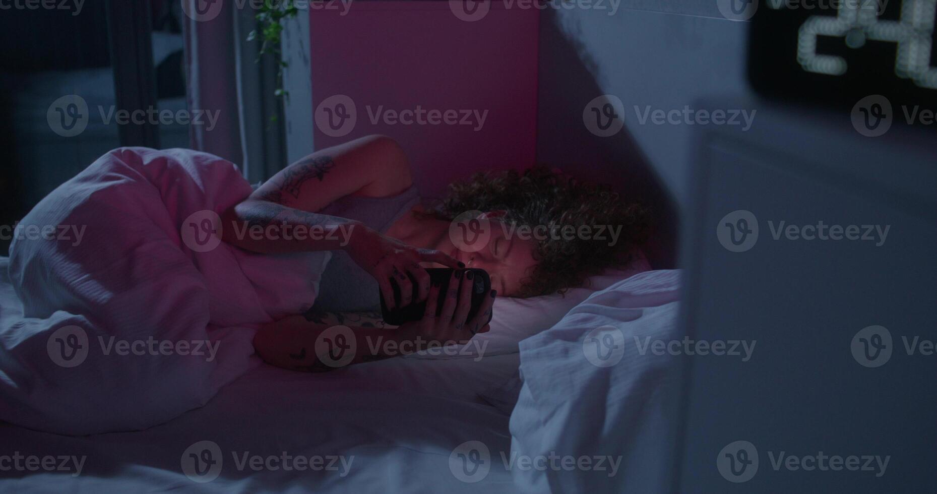 A woman uses a mobile phone lying on the bed. Dark room, home interior. The electronic clock is on the bedside table photo