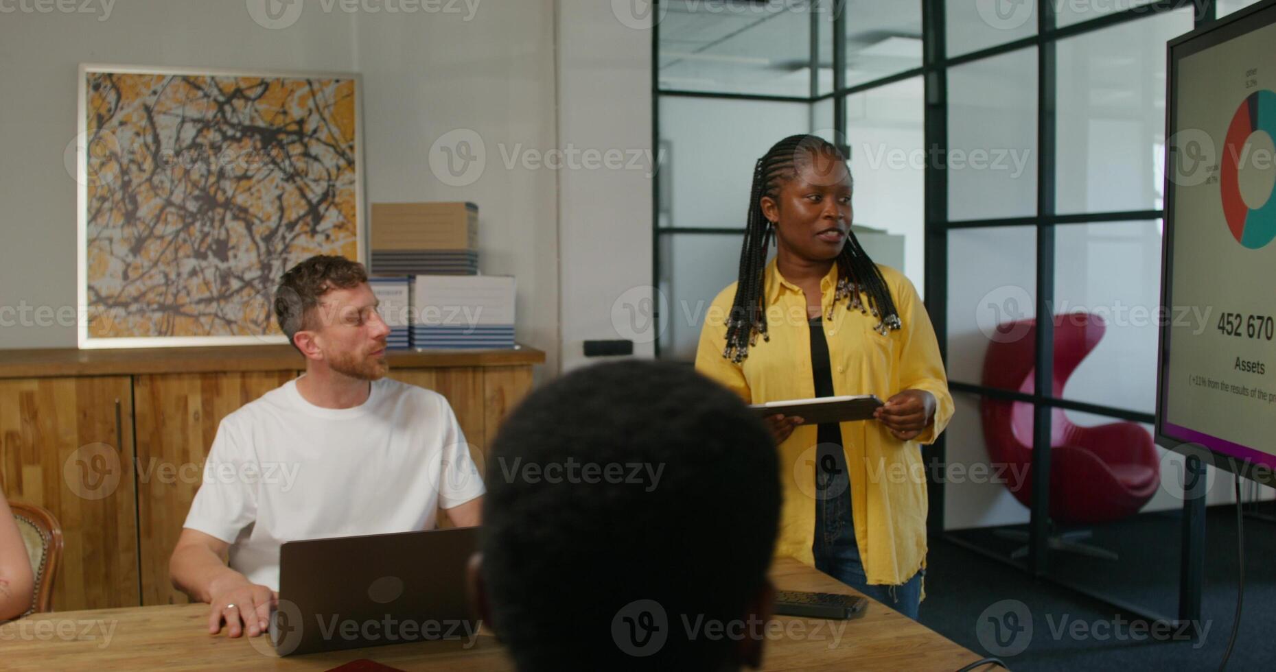 A group of young people are sitting at a table in an office meeting. A female speaker is talking standing near the screen. An open laptop is in front of a man. Daylight - office photo