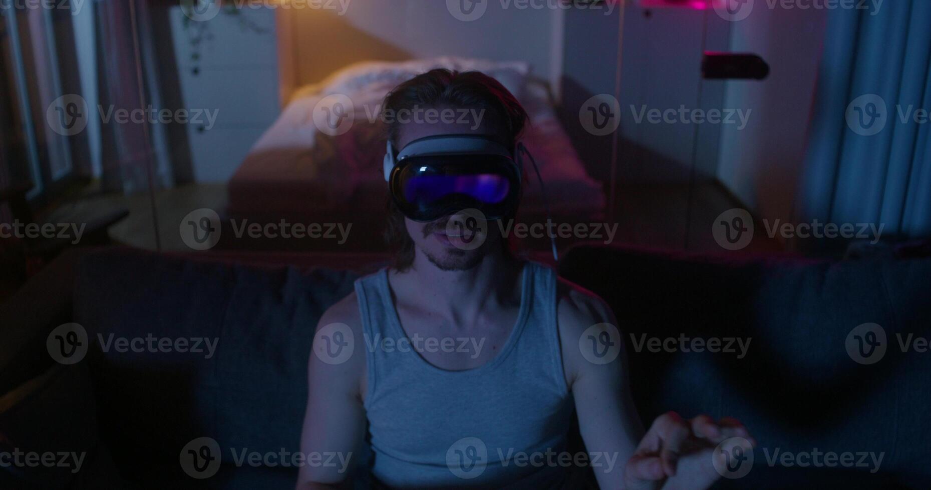 A man wearing virtual reality glasses is sitting on the couch and moving his arms. Dark room, home interior photo