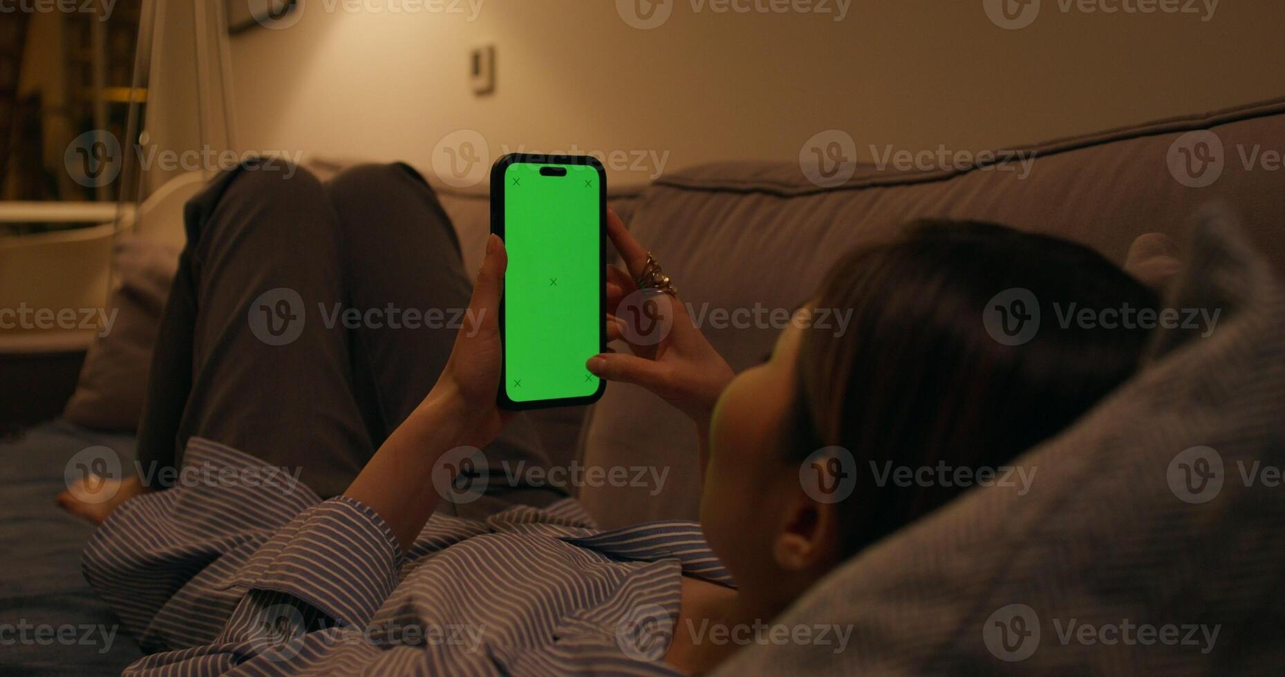A woman uses a mobile phone with a green screen lying on the couch at home photo