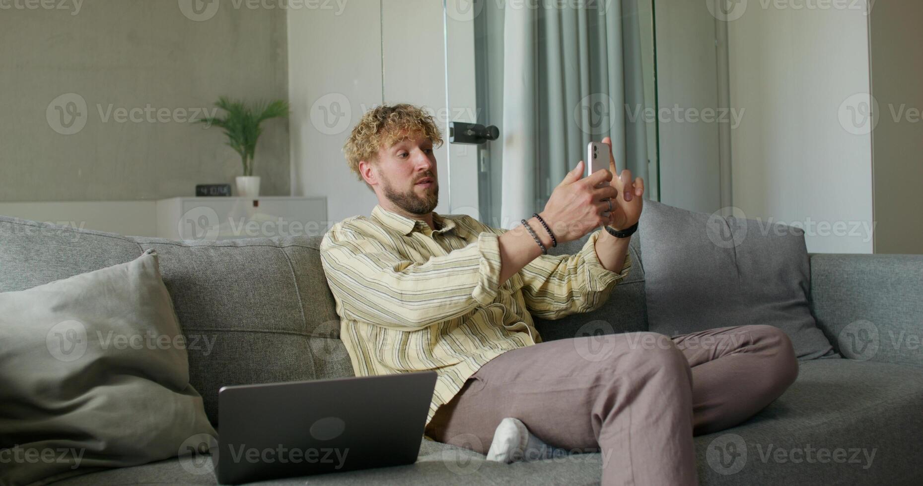 A bearded man in casual clothes is talking on a chat on a mobile phone sitting on the couch at home. The laptop is next to him. Daylight photo