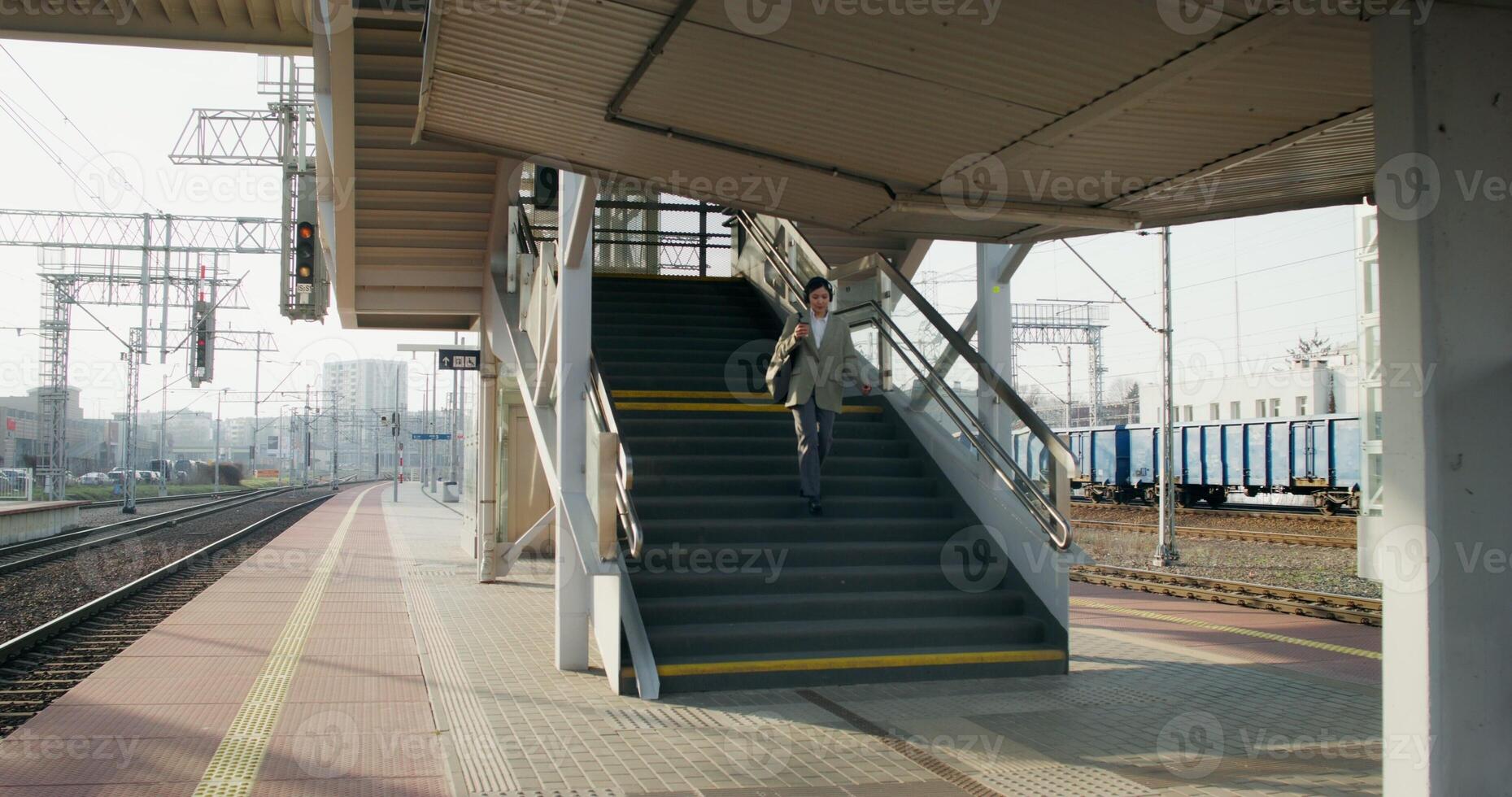 A woman in formal clothes and headphones, with a cup of coffee in her hand, descends the stairs to the platform of the railway station photo