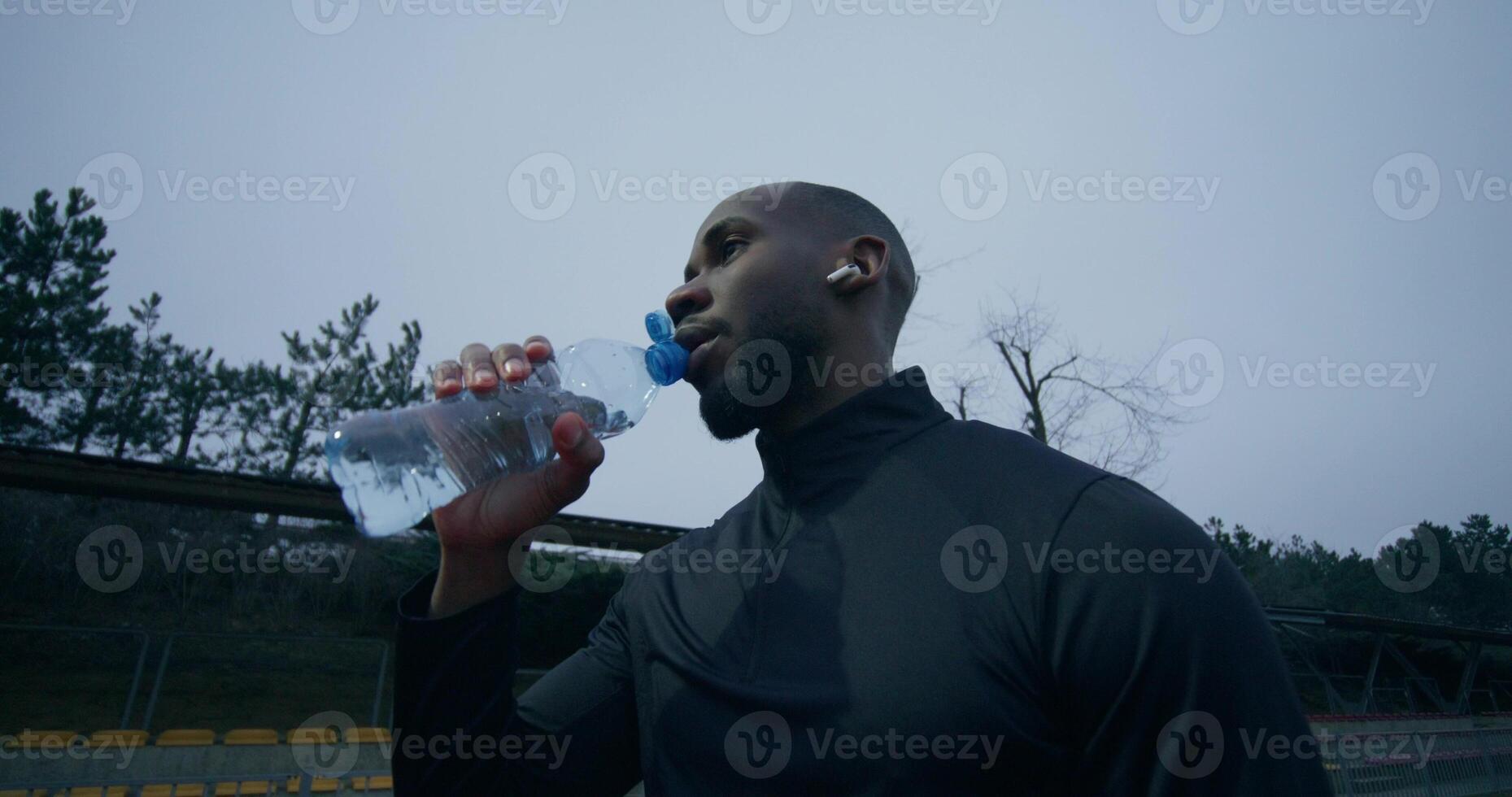 A man in sports clothes, with earphones in his ears, drinks water from a bottle standing at the stadium after jogging - drinks photo
