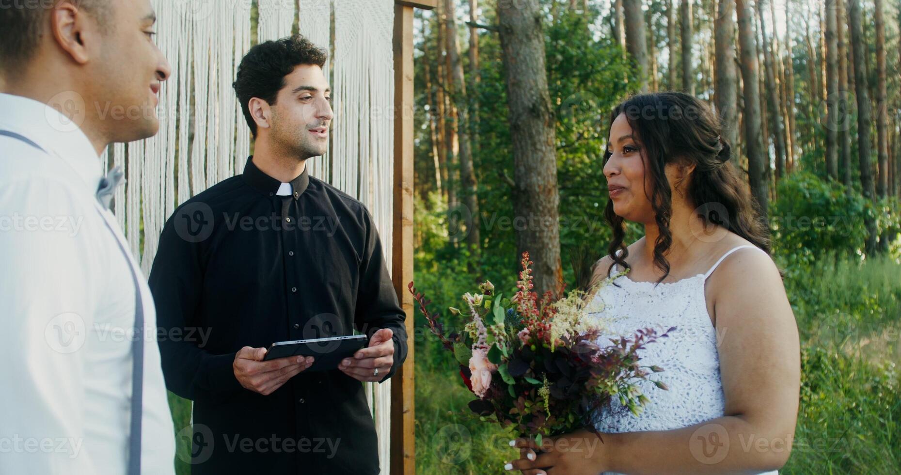 A young priest with a tablet in his hands marries an African-American couple in a beautiful pine forest near a decorated arch - marries photo