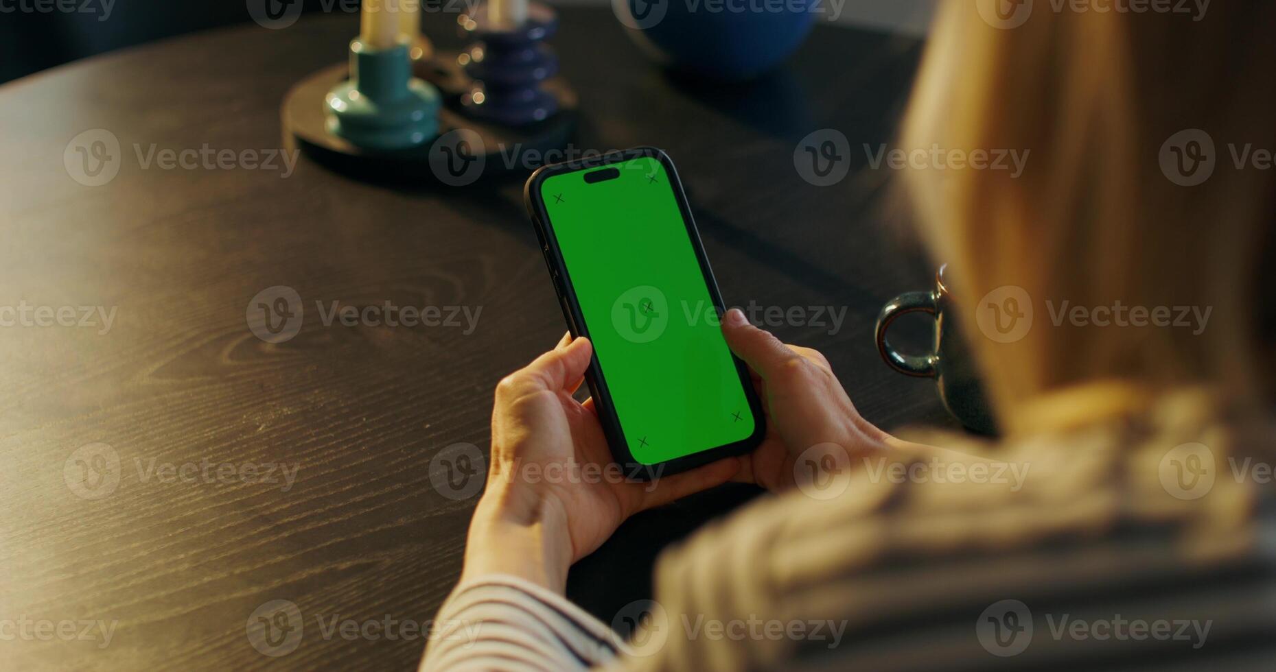 A woman uses a mobile phone with green screen sitting at a table indoors, close-up of her hands, an unrecognizable person photo