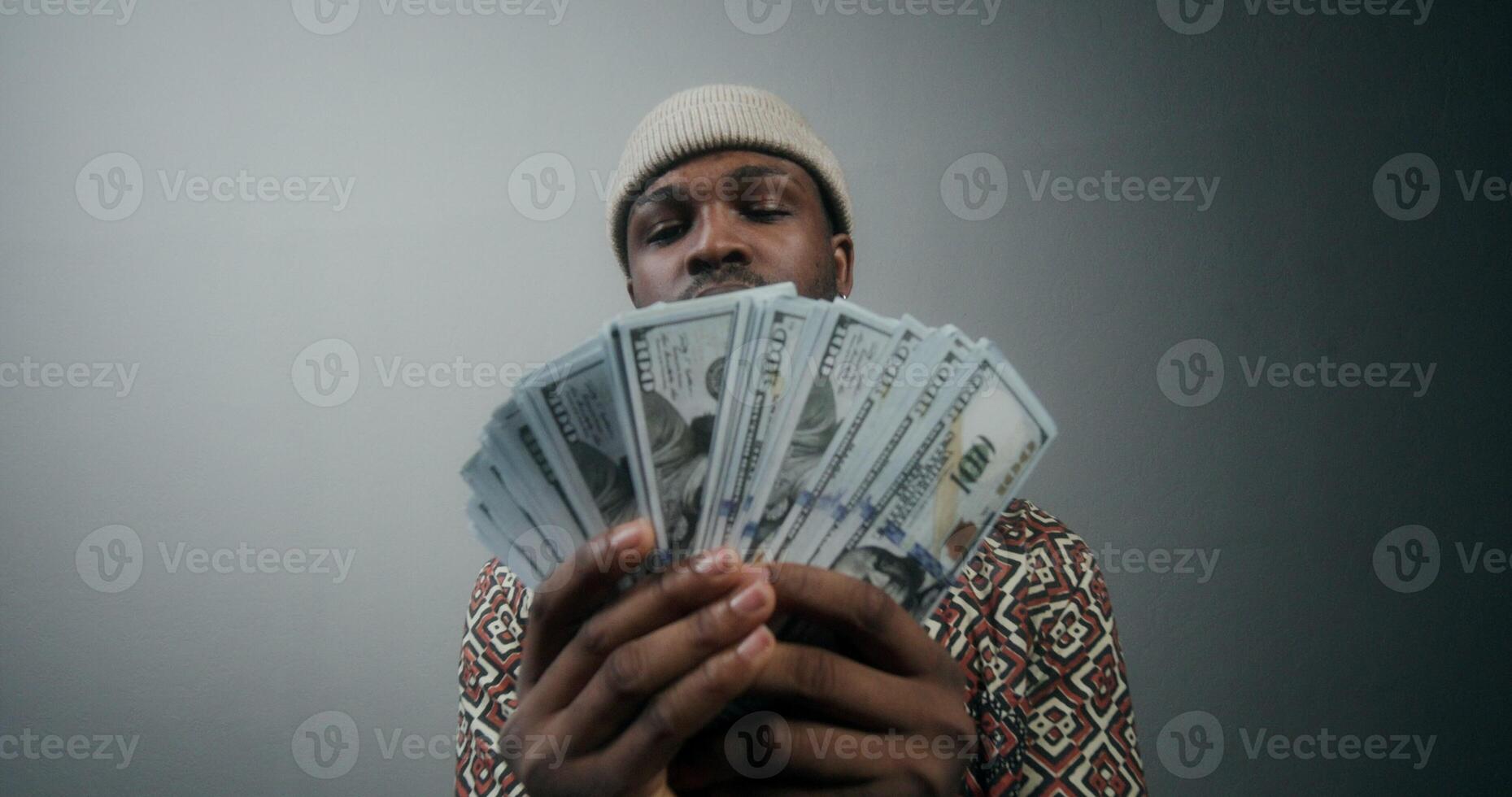A stylish bearded man in a hat tosses a stack of money and smiles at the camera standing on a gray background in the studio photo