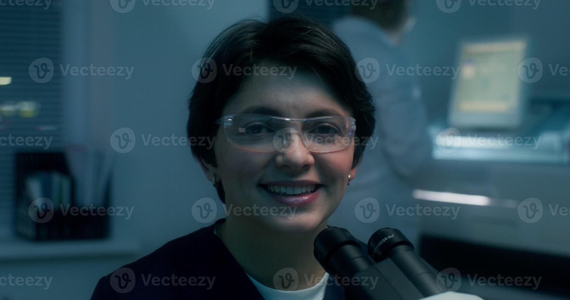 A woman in a lab coat and safety glasses smiles looking at the camera sitting in front of a microscope in a medical laboratory - microscope photo