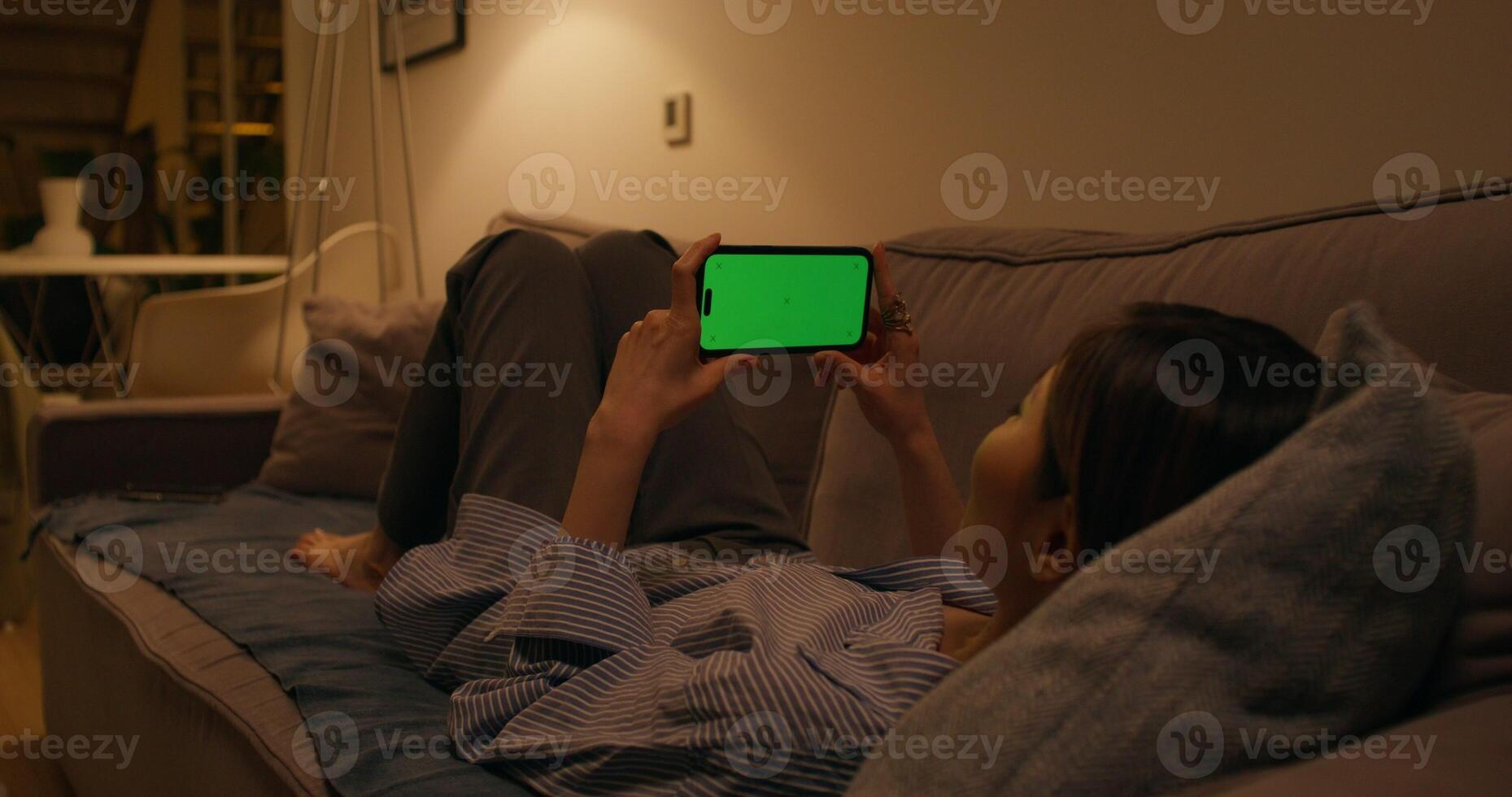 A woman uses a mobile phone with a green screen lying on the couch at home, dimmed evening light - mobile photo