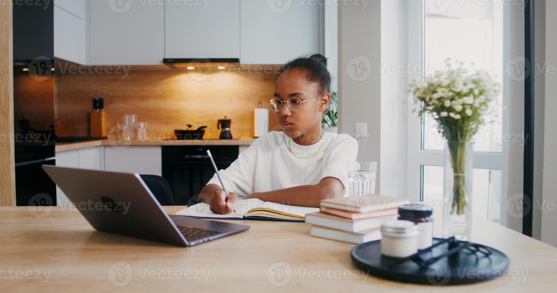 The girl does her homework at home, she uses a laptop and makes notes in a notebook while sitting at the kitchen table - sitting photo