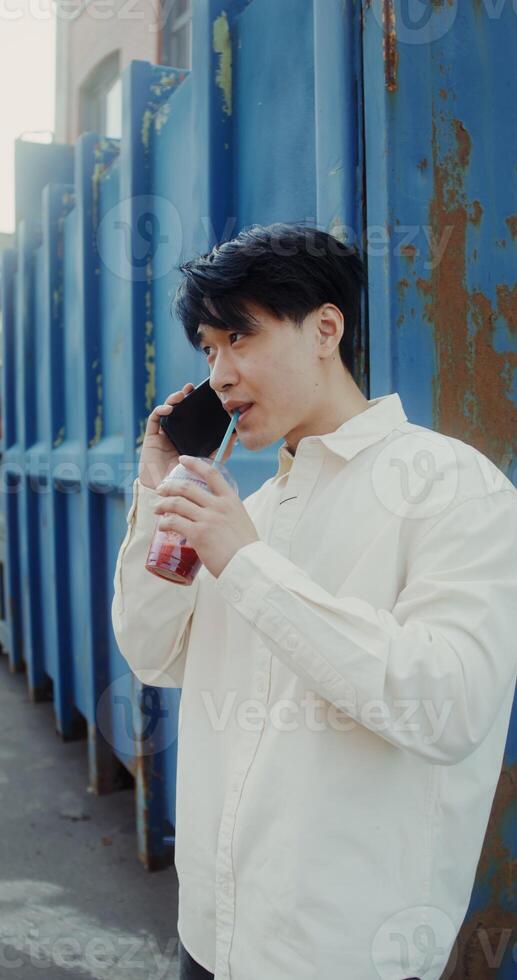A young man of Chinese appearance is talking on a mobile phone, and drinking juice from a disposable cup with a straw standing near a metal wall outdoors - talking photo