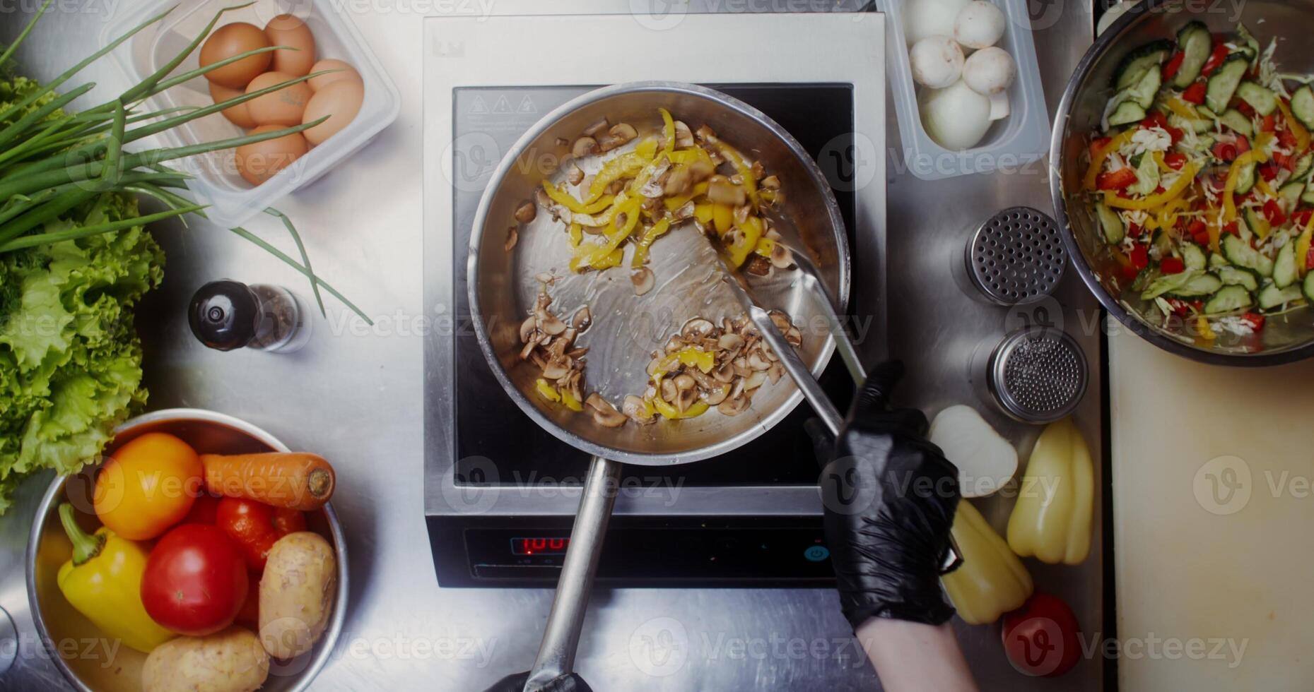 A woman in disposable black gloves mixes mushrooms with pepper in a frying pan with tongs, top view, close photo