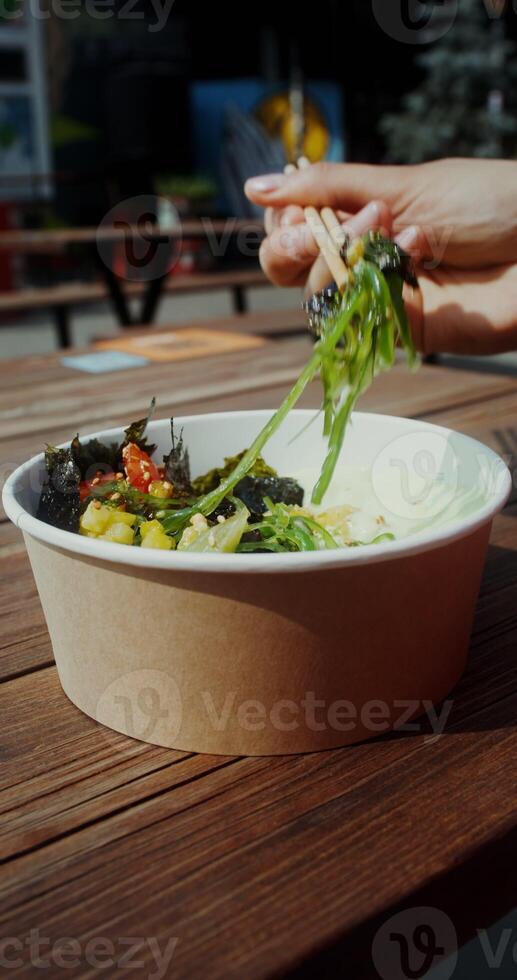 A young woman eats Asian food out of a box using chopsticks while sitting at a table on the cafe terrace, hands close-up, no face - sitting photo
