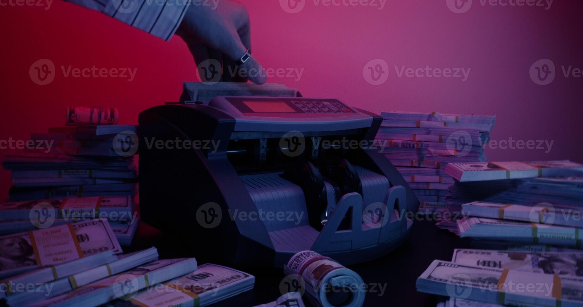 A man's hand with a ring inserts a stack of hundred-dollar bills into the counting machine and it starts counting. Bundles of money are lying around on the table photo