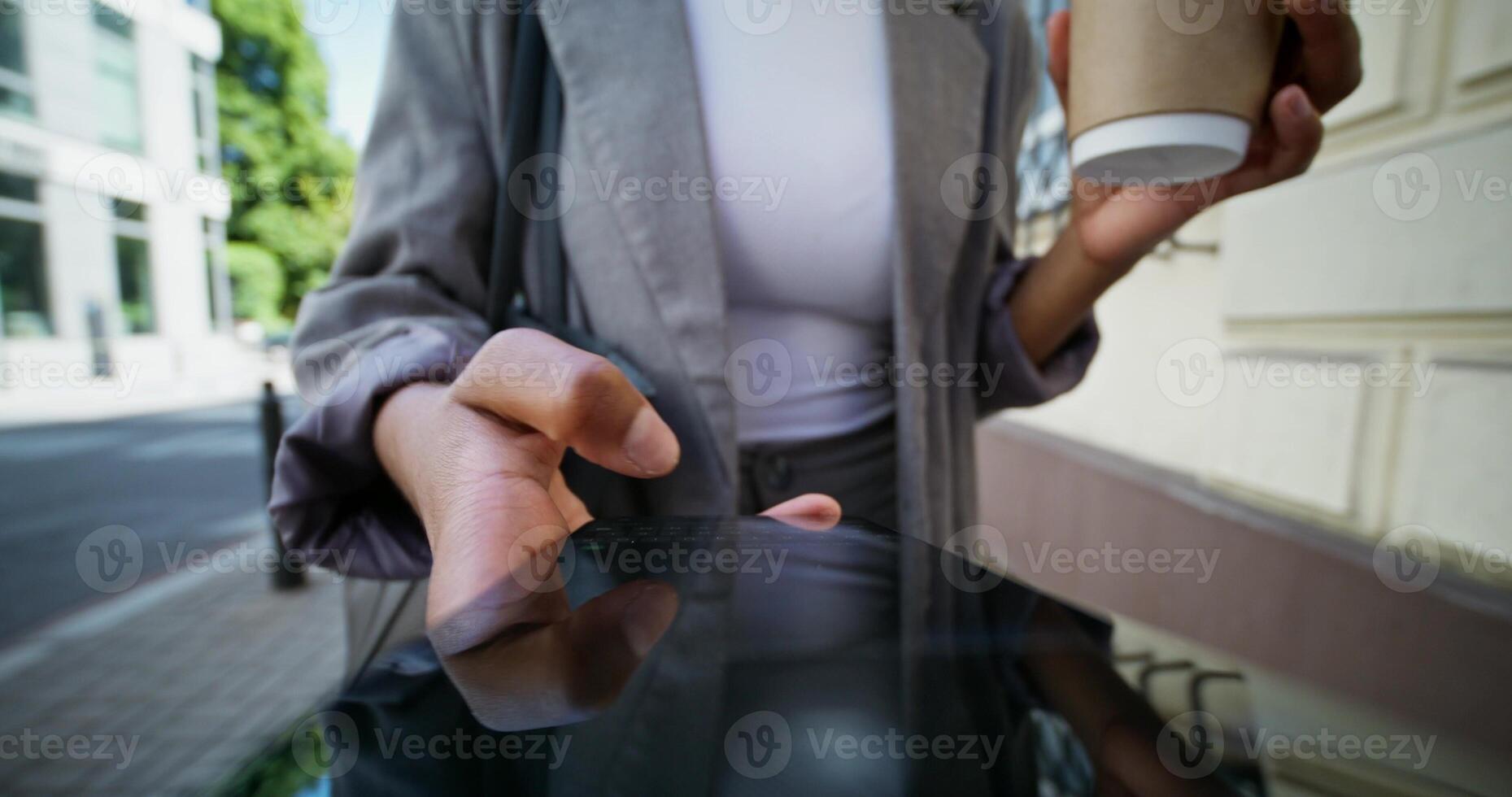 A woman in formal clothes uses a mobile phone walking along a city street. She holds a disposable cup of coffee in her hand. Close-up of a mobile phone screen, an unrecognizable person - mobile photo