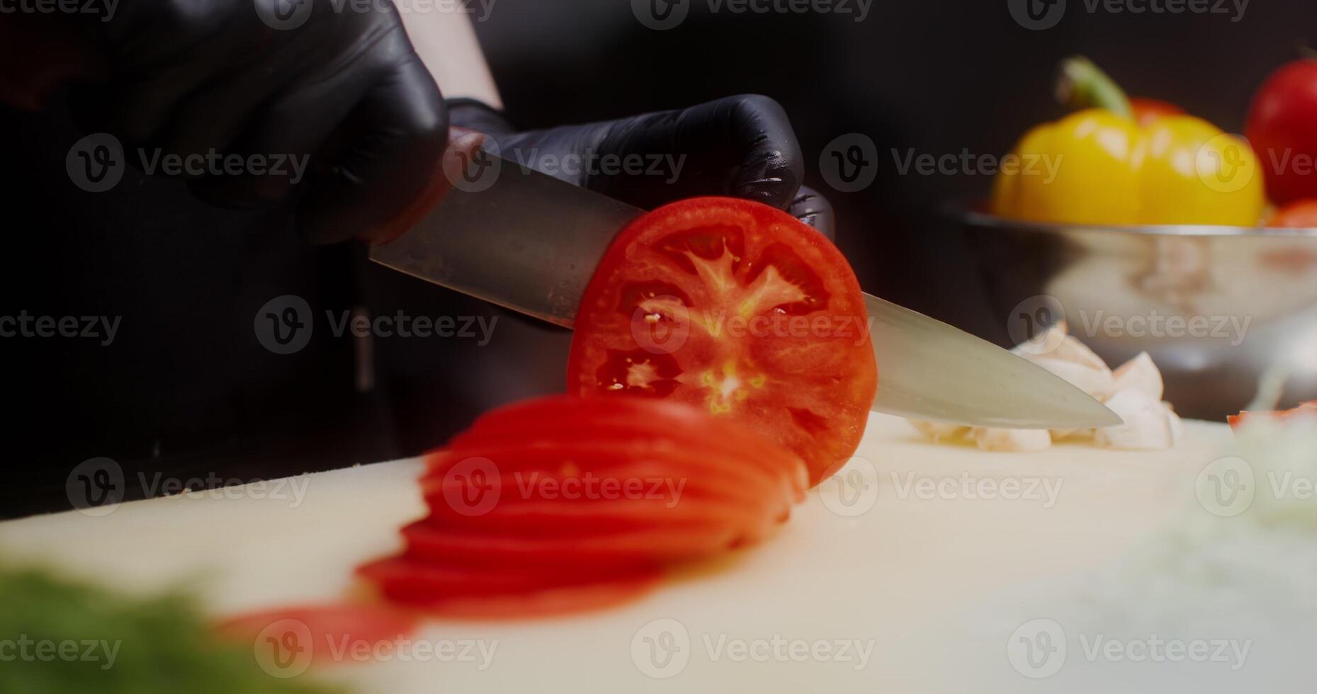 A woman in a chef's uniform with disposable black gloves on her hands cuts tomato standing in the restaurant kitchen. Close-up of a woman's hands, no face - tomato photo