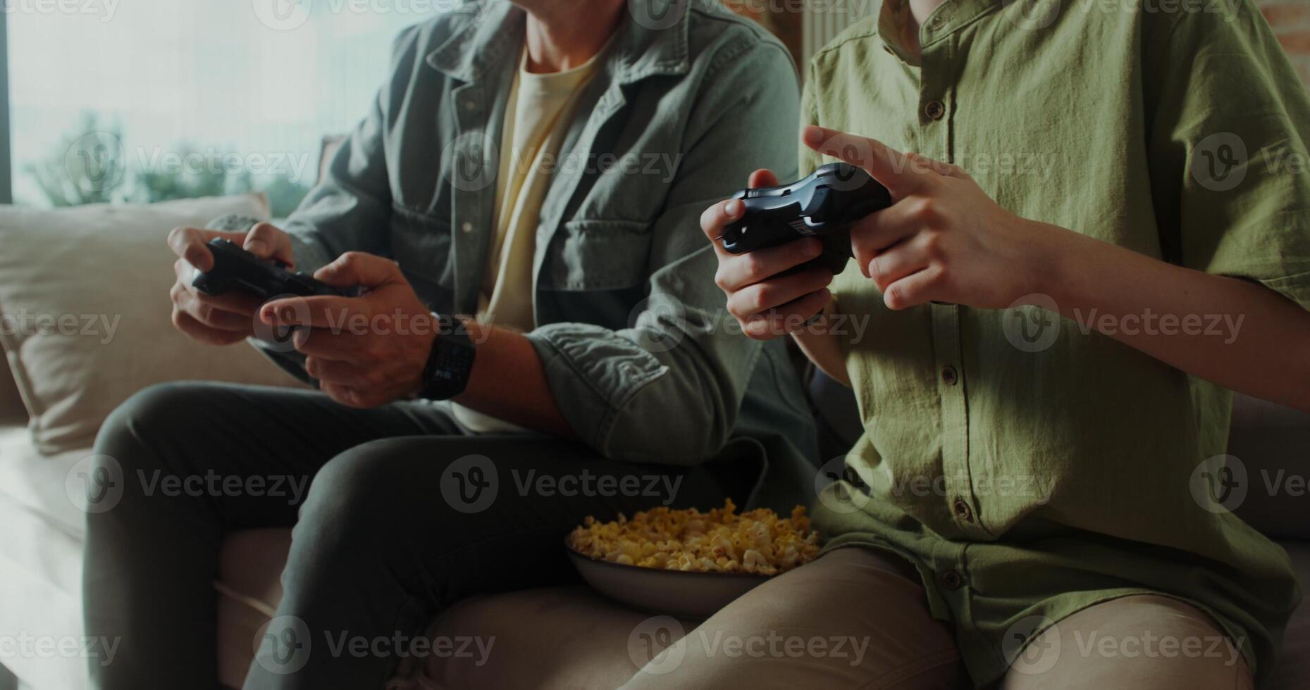 Father and son play games with joysticks and eat popcorn sitting on the couch at home, happy childhood - popcorn photo