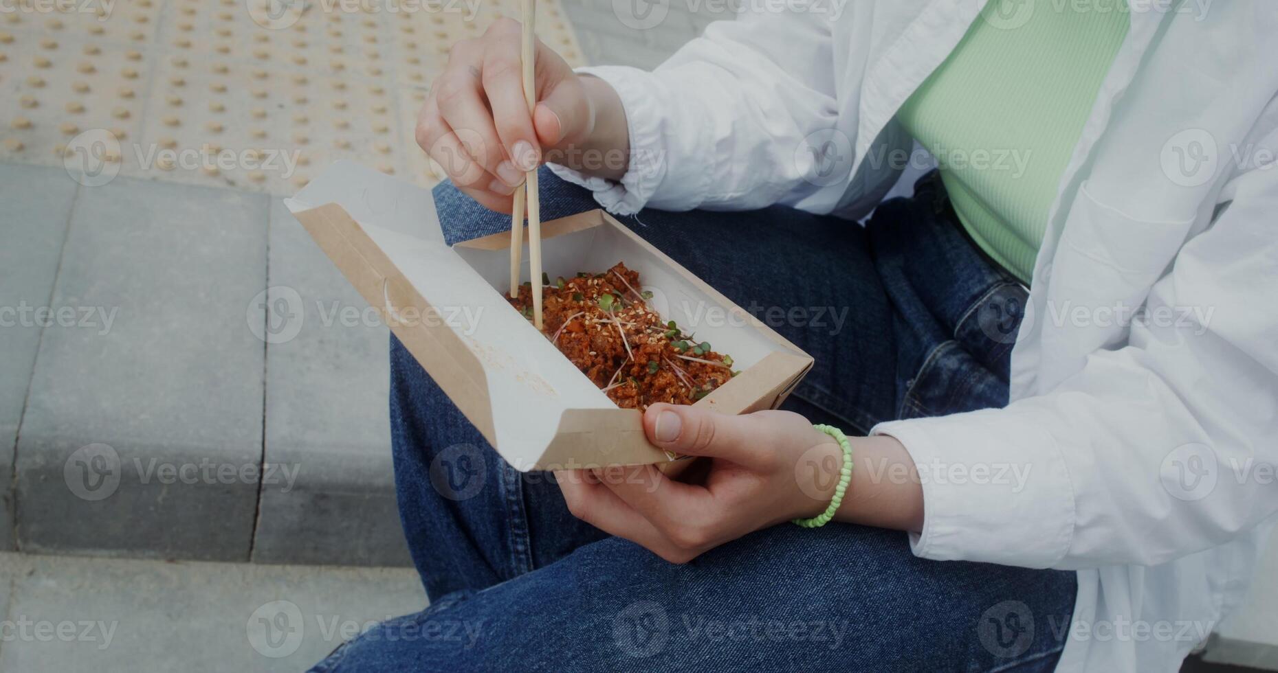 A woman eats noodles out of a box using chopsticks sitting outdoors, close-up of a woman's hands, no face - sitting photo