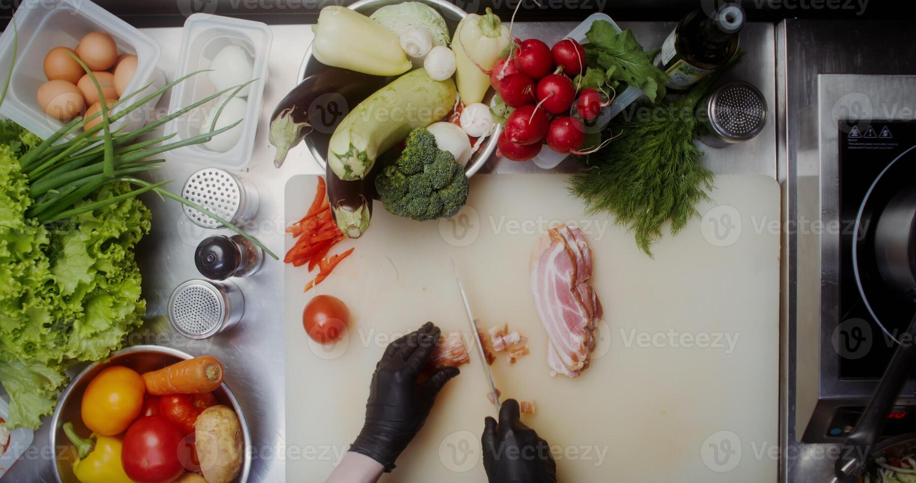 A woman in disposable black gloves cuts bacon while working at a table full of vegetables, close-up of her hands, top view - working photo