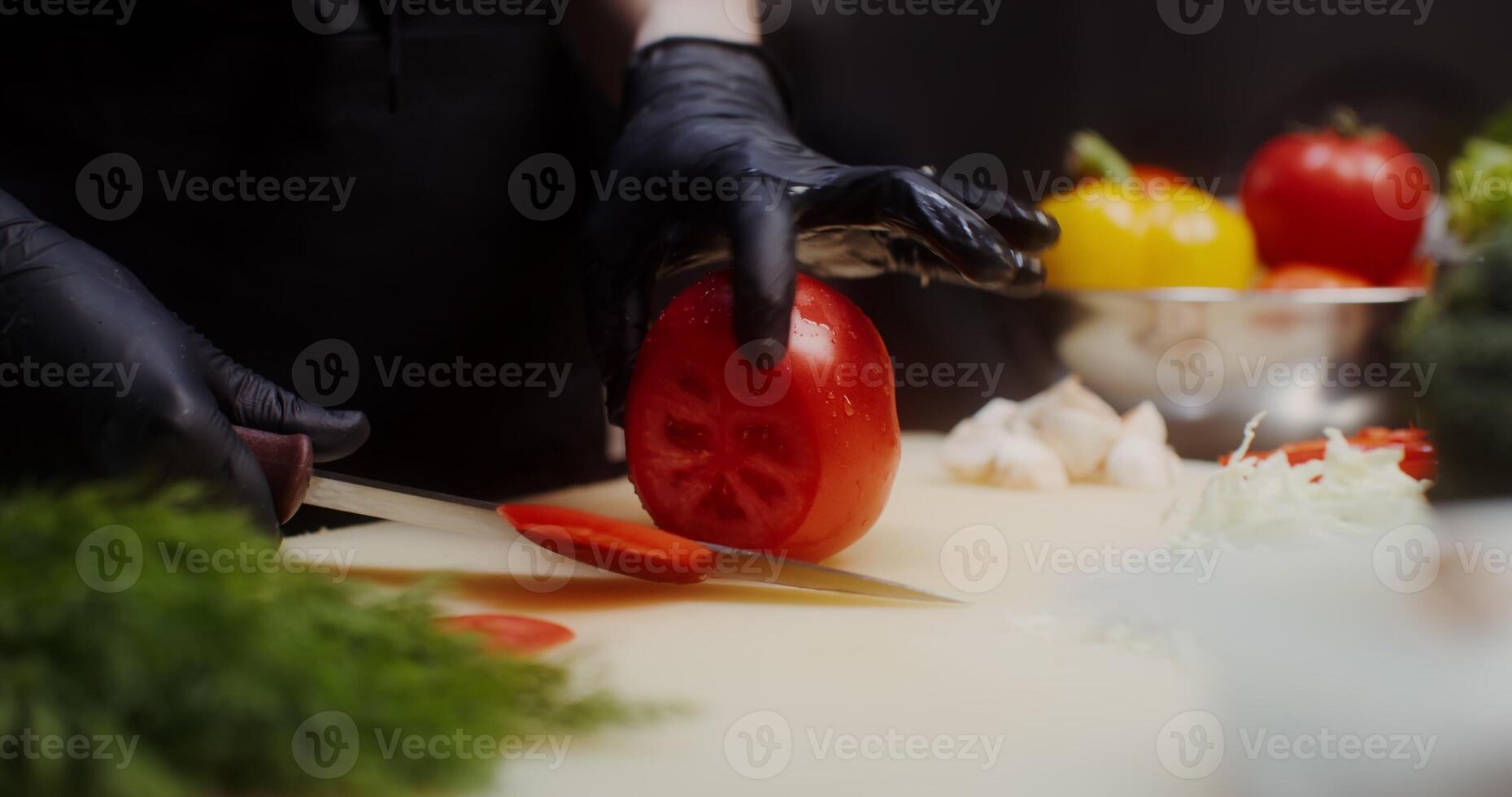 A woman in a chef's uniform with disposable black gloves on her hands cuts tomato standing in the restaurant kitchen. Close-up of a woman's hands, no face - gloves photo