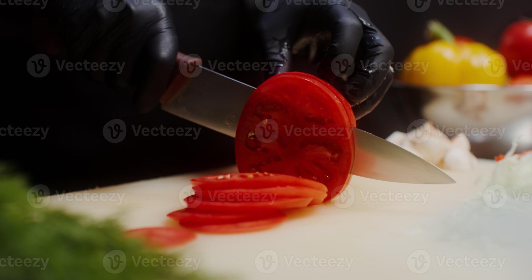 A woman in a chef's uniform with disposable black gloves on her hands cuts tomato standing in the restaurant kitchen. Close-up of a woman's hands, no face - standing photo