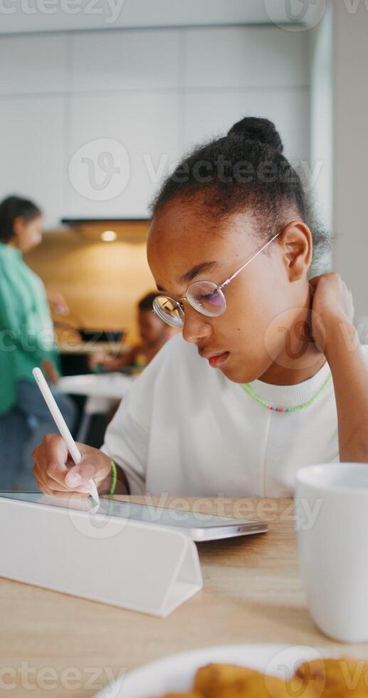 A girl draws with a stylus on a tablet while sitting at the kitchen table at home. Her family in the background is out of focus - background photo