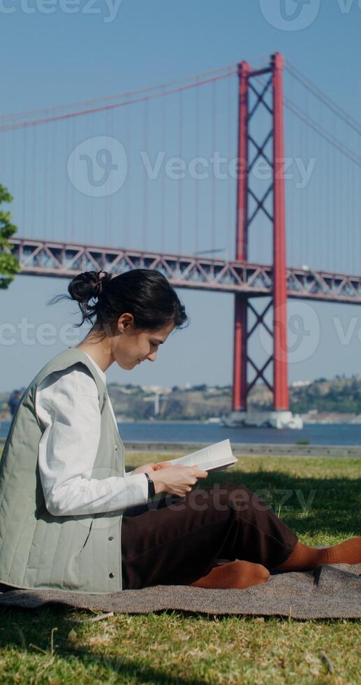 A young beautiful woman is reading a book sitting on a blanket near the city embankment with a red bridge over the river - sitting photo