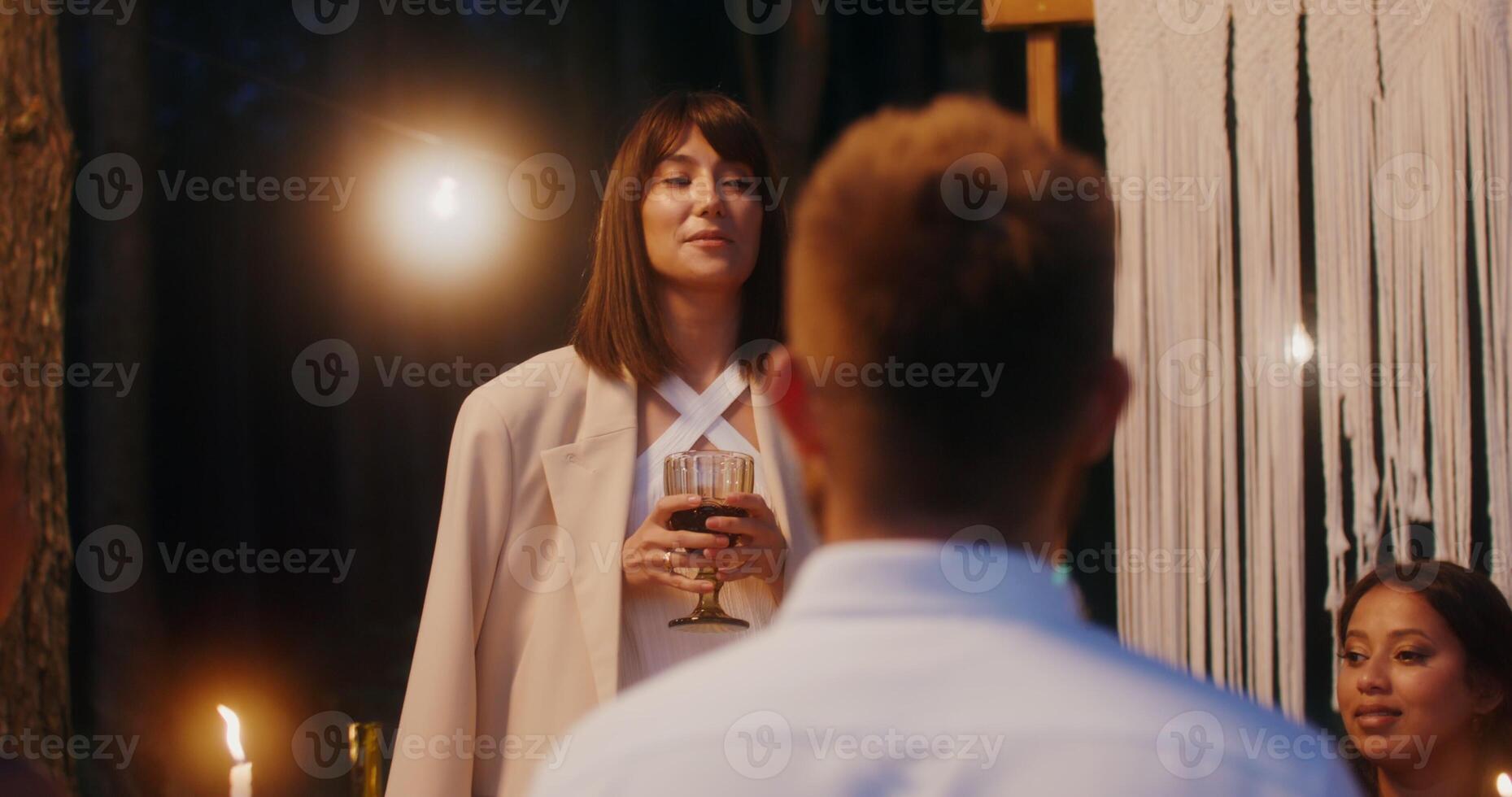 Guests smile sitting at a beautifully served festive table outdoors at night under the light bulbs, a woman stands saying congratulations - beautifully photo