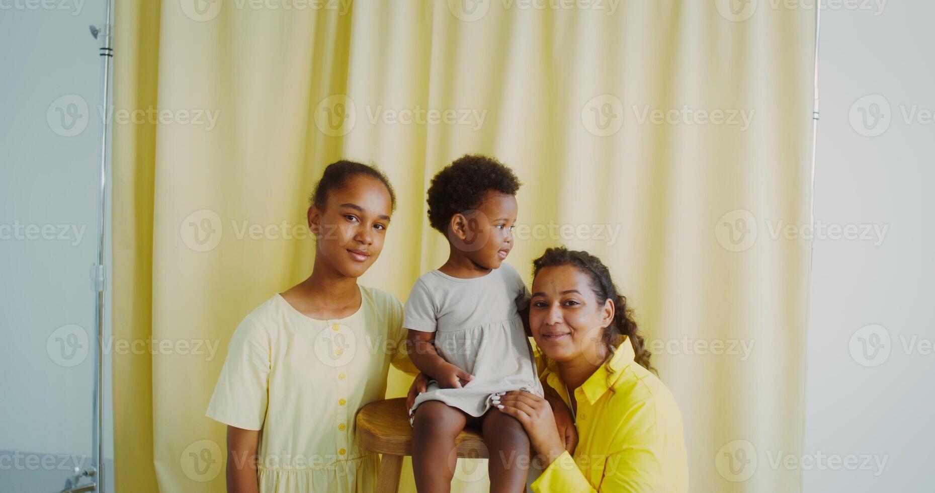 A mother with two children smiles while looking at the camera posing in the studio. The baby is sitting on a high chair photo