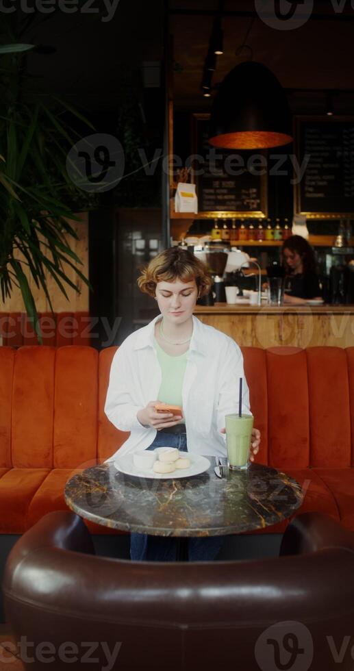 A woman in casual clothes uses a mobile phone and drinks a cocktail while having breakfast in a cafe photo