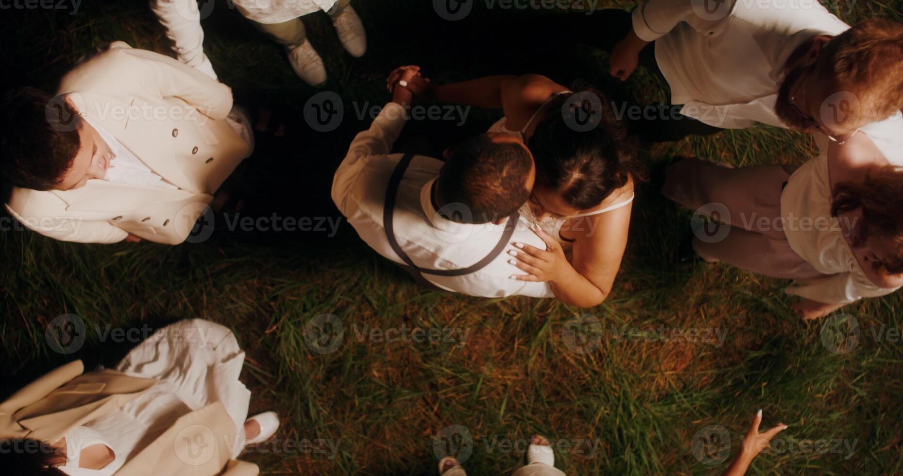 Elegant guests smile and dance leading a round dance around the bride and groom at an outdoor party at night, top view - leading photo