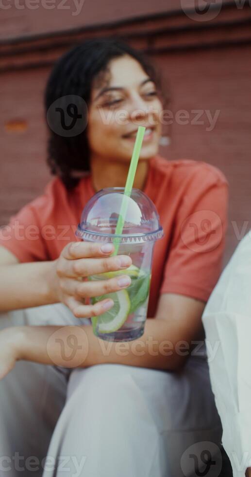 A woman drinks juice from a disposable cup with a straw while sitting on a city street, and talks to a person sitting next to her - sitting photo