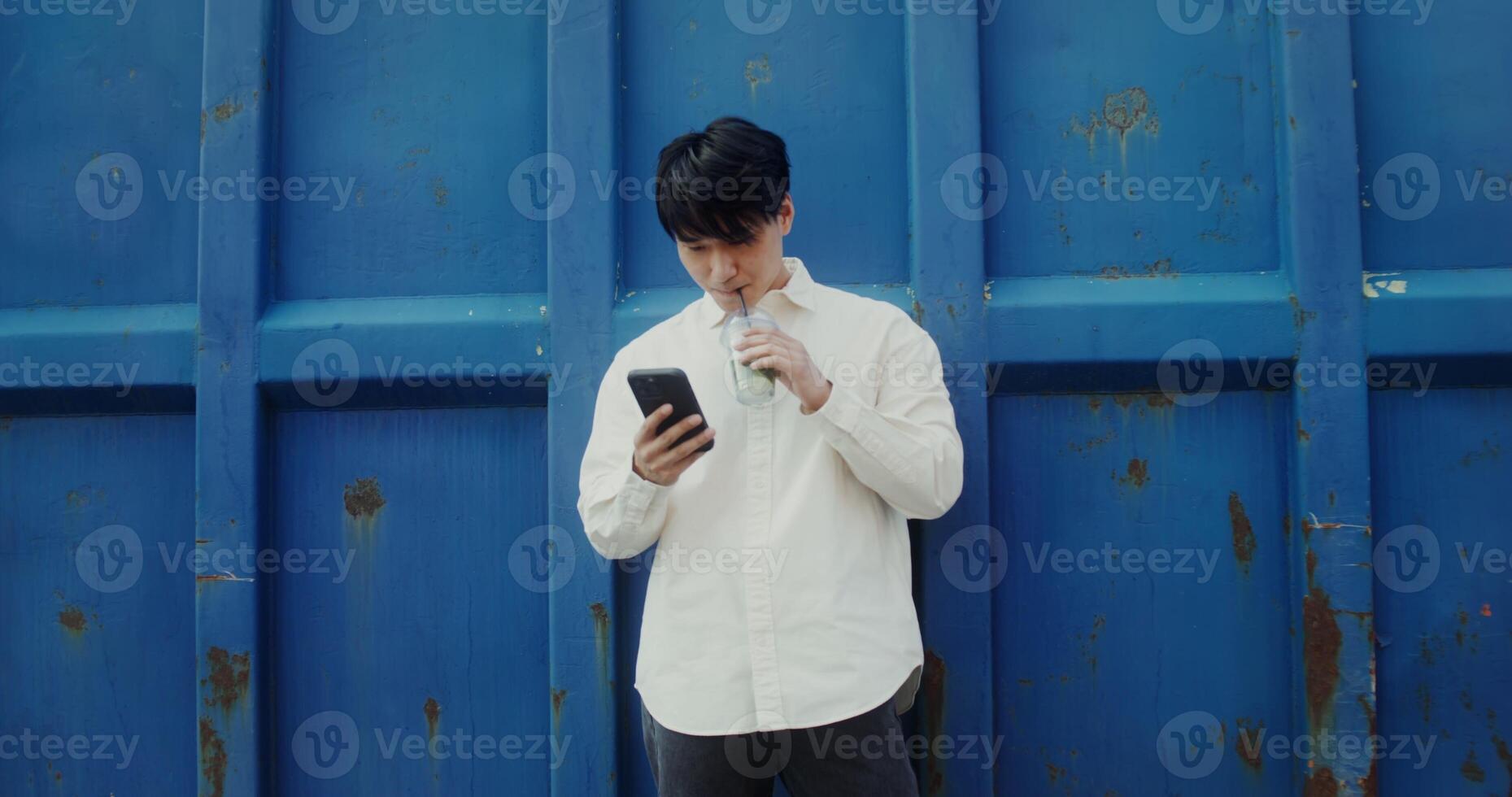 A young man of Chinese appearance uses a mobile phone and, drinks mojito from a disposable cup with a straw standing near a metal wall outdoors - drinks photo