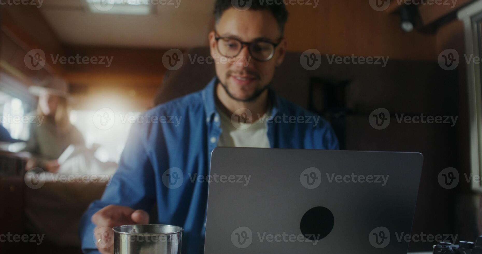 A young man uses a laptop and drinks a hot drink from a metal mug while sitting at a table in a van. A woman reads a book in the background - laptop photo