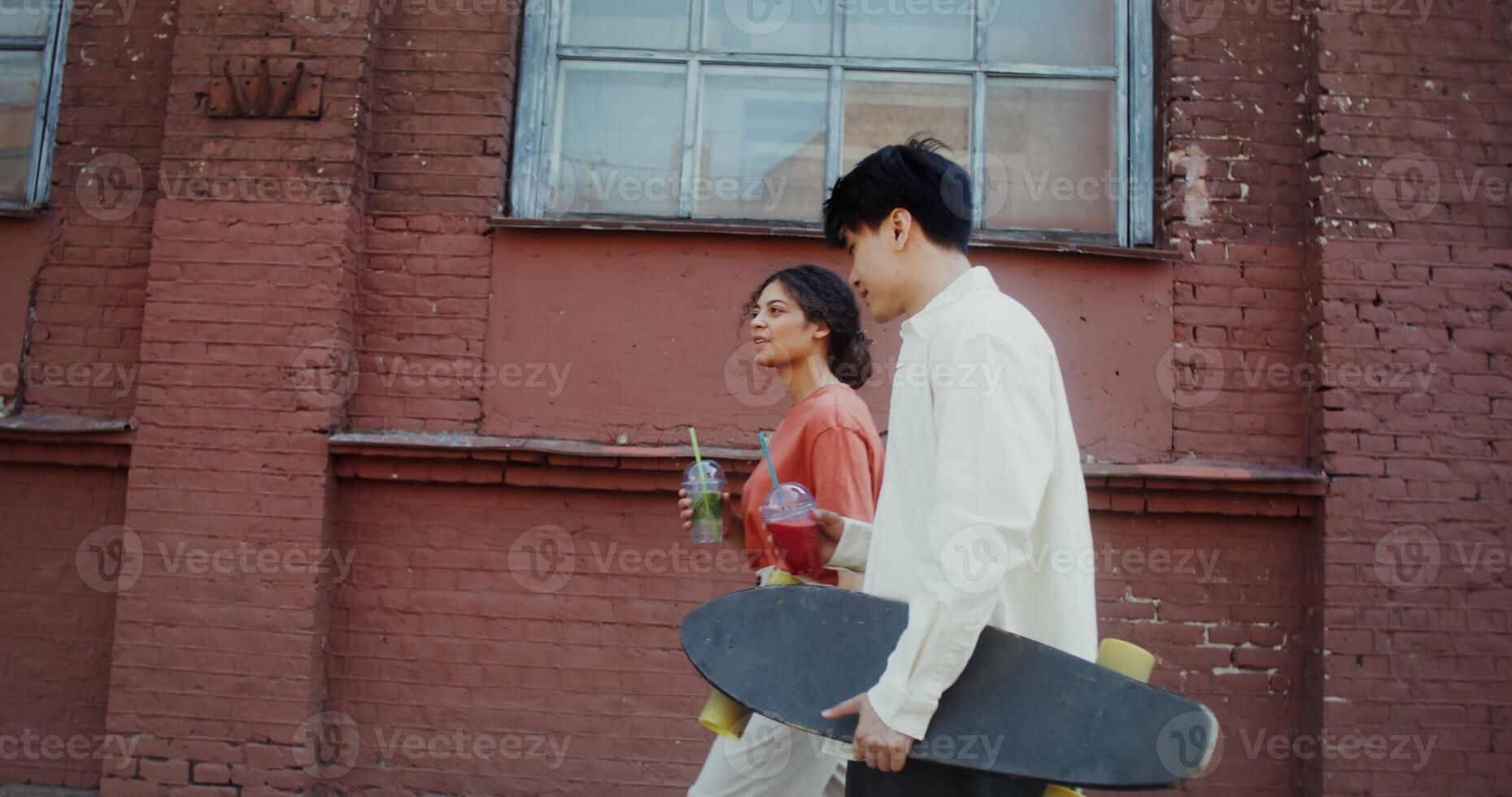 A young man and a woman smile, talk and drink lemonade while walking around the city. A man holds a longboard in his hands - around photo