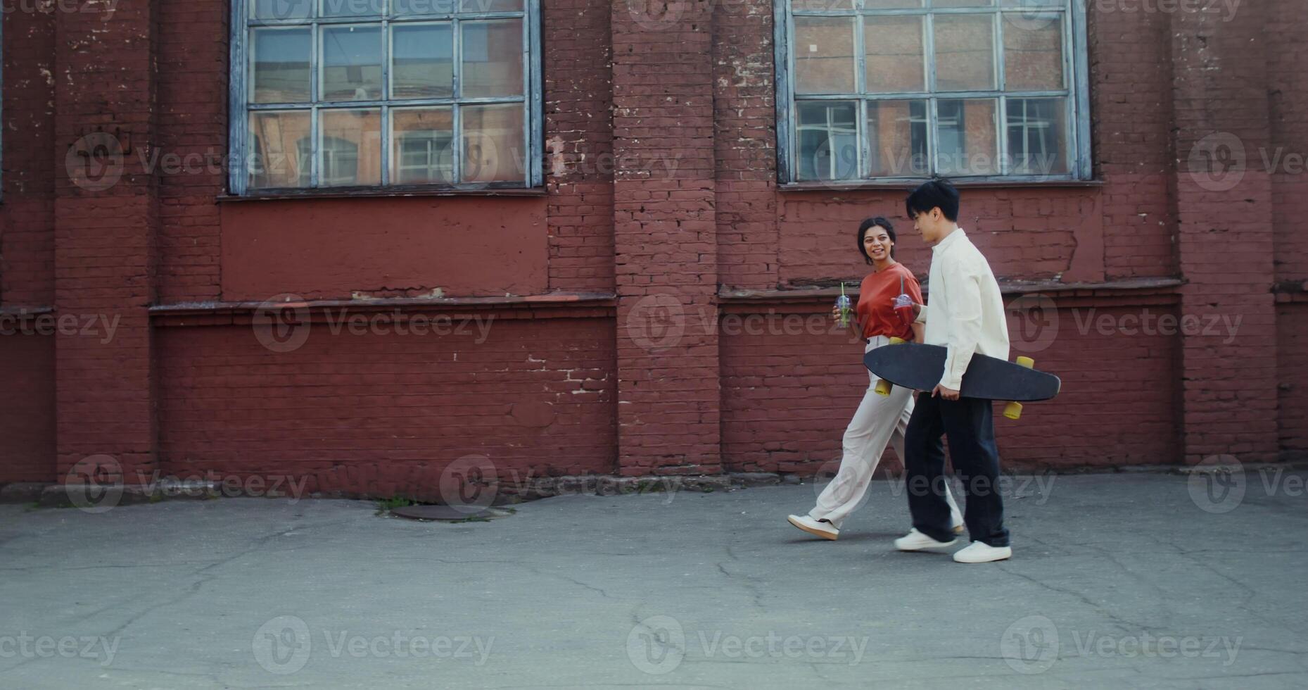 A young man and a woman smile, talk and drink lemonade while walking around the city. A man holds a longboard in his hands photo