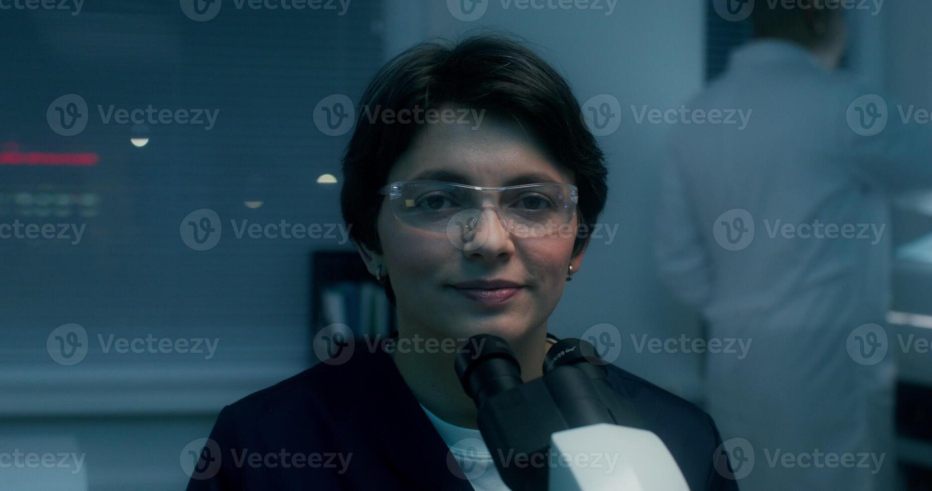 A woman in a lab coat and safety glasses smiles looking at the camera sitting in front of a microscope in a medical laboratory - camera photo
