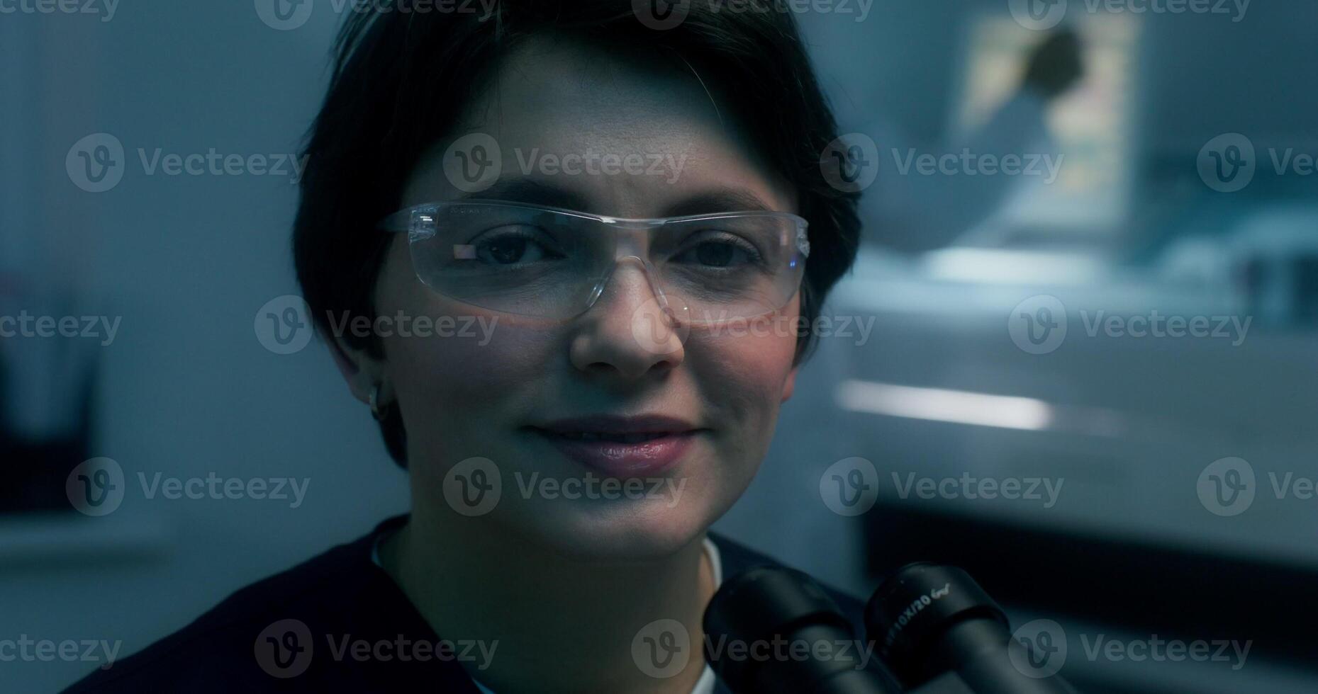 A woman in a lab coat and safety glasses smiles looking at the camera sitting in front of a microscope in a medical laboratory - sitting photo